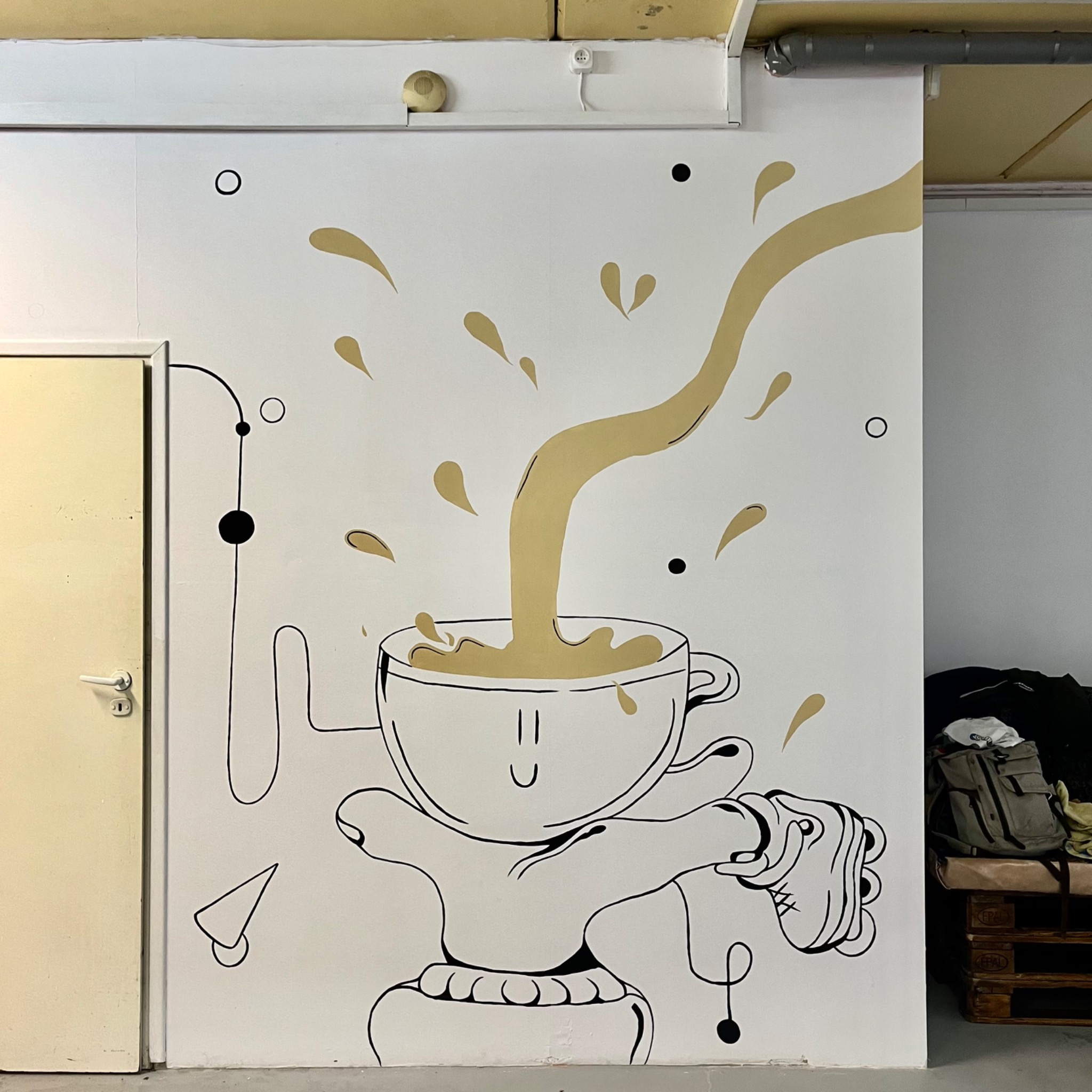 Abstrakcyjne malowidło ścienne w Katowicach przedstawiające filiżankę kawy z wylewającym się napojem, wykonane czarną i złotą farbą na białej ścianie, obok kremowych drzwi.