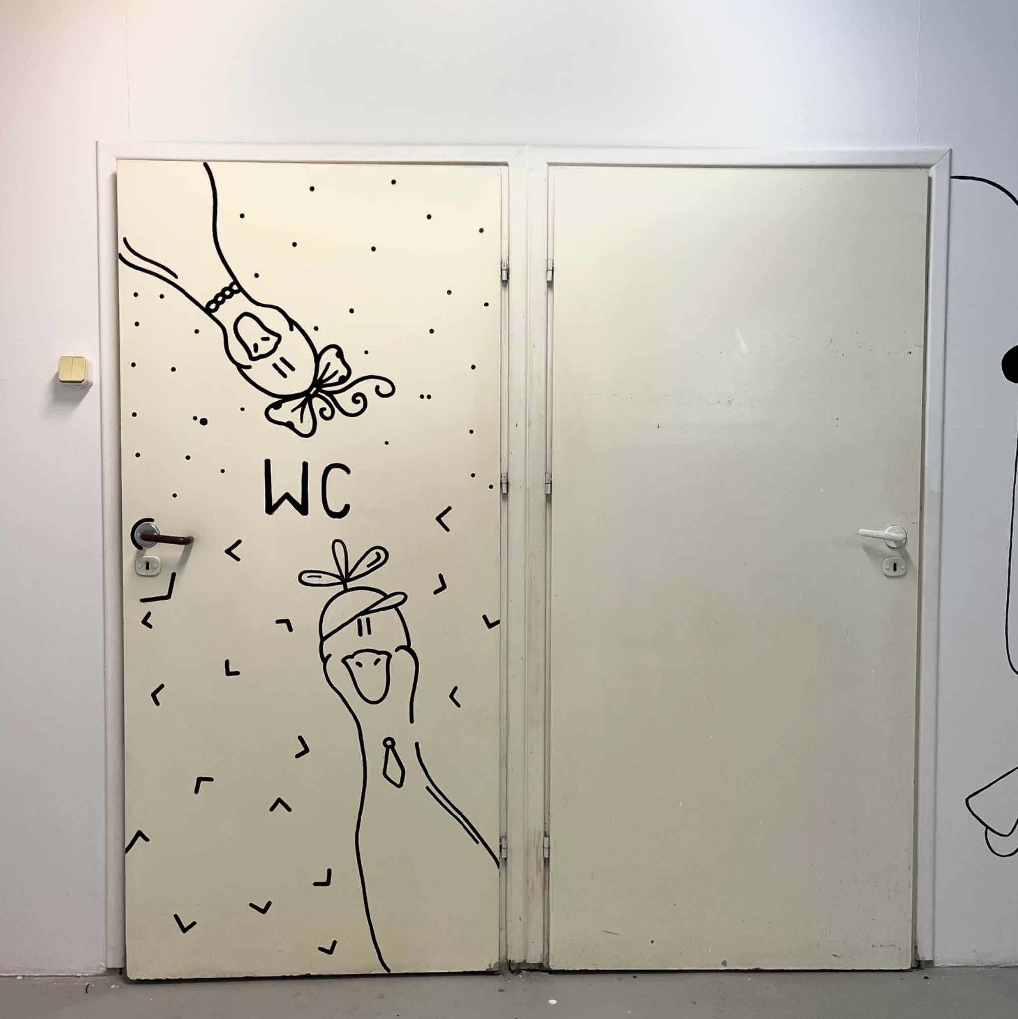 Drzwi do toalety z ręcznie malowanymi, czarnymi rysunkami postaci i napisem WC, w stylu line art.