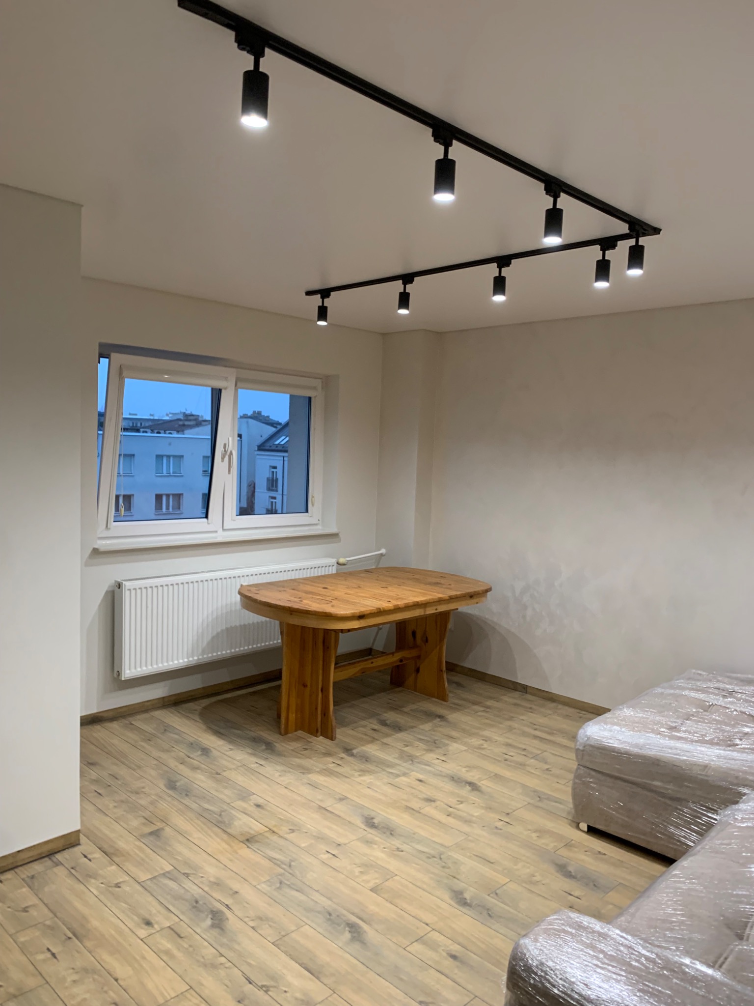 Jasne wnętrze pokoju z drewnianym stołem, panelami podłogowymi i nowoczesnym oświetleniem szynowym; sofa owinięta folią ochronną.