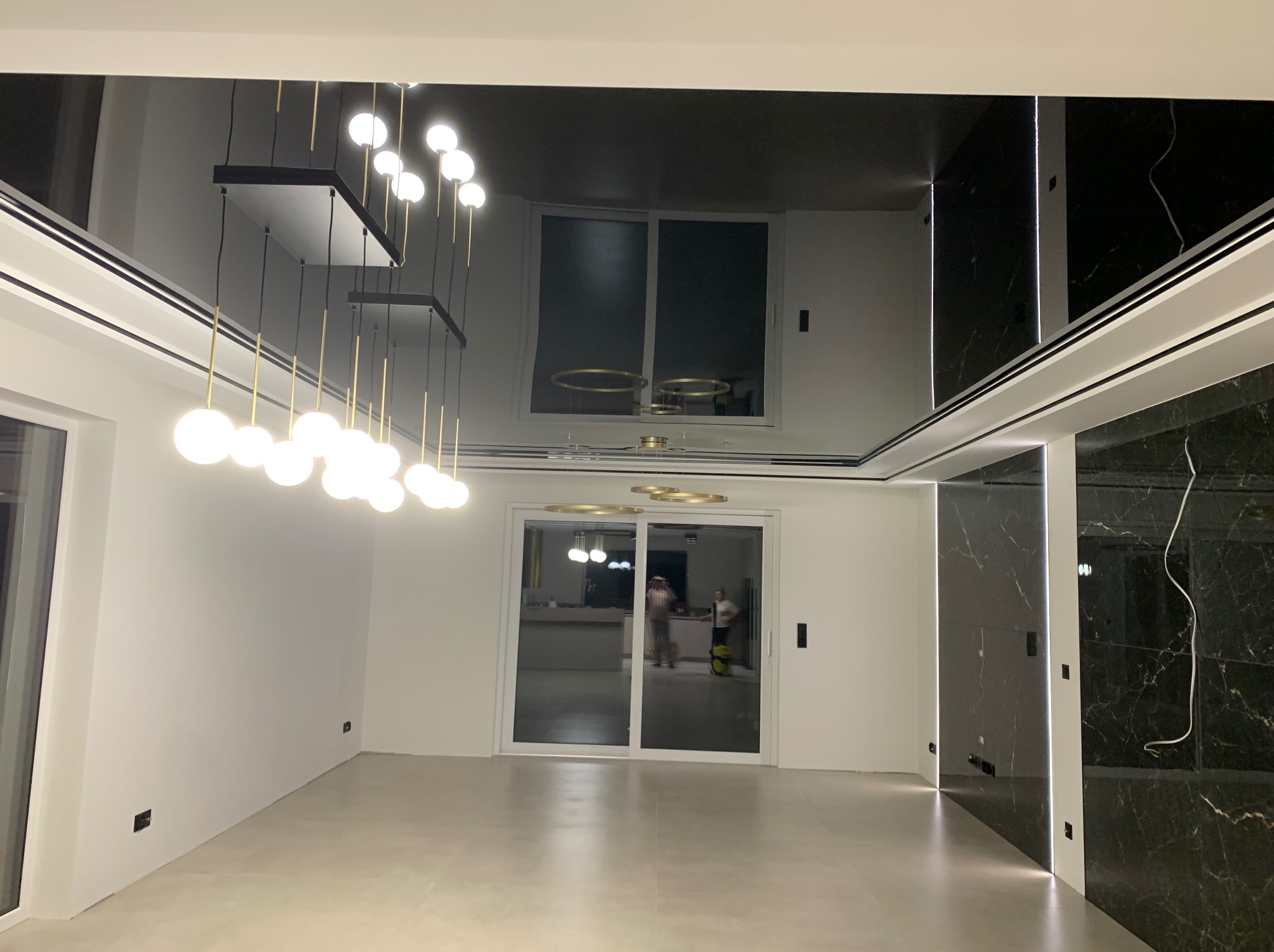 Nowoczesne wnętrze z czarnym lustrzanym sufitem, wiszącymi lampami z kulistymi kloszami i ścianami wykończonymi białymi panelami oraz czarnym marmurem z podświetleniem LED, widok na przesuwne drzwi...