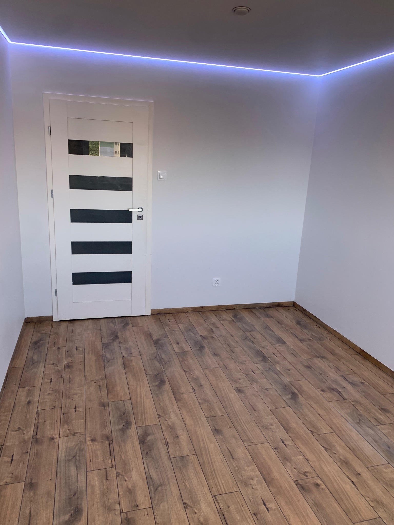 Pokój z ułożonymi panelami podłogowymi o wzorze drewna, białe ściany, białe drzwi z czarnymi poziomymi paskami i oświetleniem LED przy suficie.