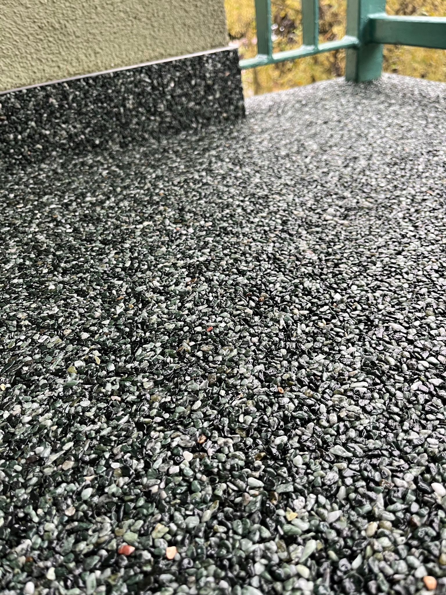 Szczegółowe ujęcie posadzki żywicznej na balkonie z zieloną balustradą w tle, widoczna struktura kamyczków i fragment ściany.