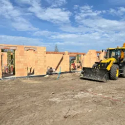 Wznoszenie ścian domu z czerwonej cegły ceramicznej. Żółta koparko-ładowarka JCB na placu budowy. Błękitne niebo z obłokami w tle.