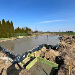 Wylany fundament z wystającymi prętami zbrojeniowymi, zabezpieczony czarną folią, otoczony drewnianym szalunkiem. W tle rząd drzew i błękitne niebo.