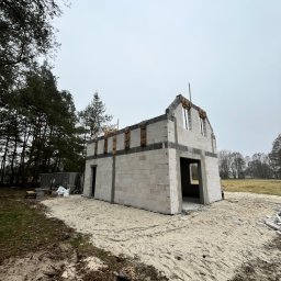 Pro House - Surowy stan domu murowanego z betonu komórkowego, widok z zewnątrz na tle drzew i nieba. Widoczne otwory okienne i drzwiowe, teren piaszczysty.