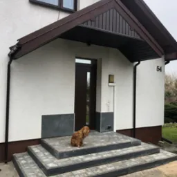 Wejście do domu z trzema stopniami wyłożonymi szarą kostką, brązowy daszek nad drzwiami, pies rasy cocker spaniel siedzi na schodach, numer domu 54.