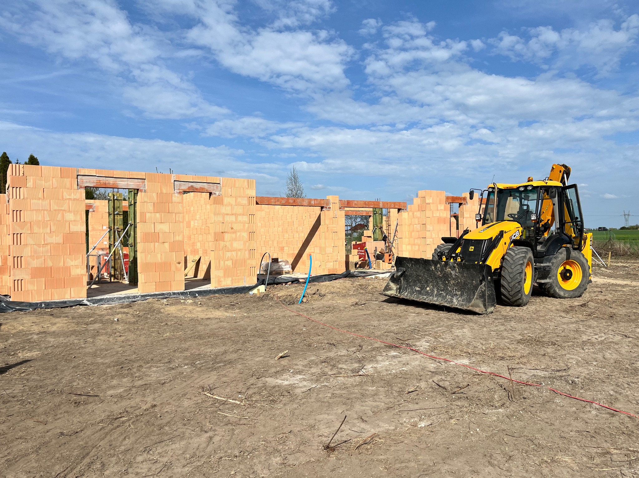 Wznoszenie ścian domu z czerwonej cegły ceramicznej. Żółta koparko-ładowarka JCB na placu budowy. Błękitne niebo z obłokami w tle.