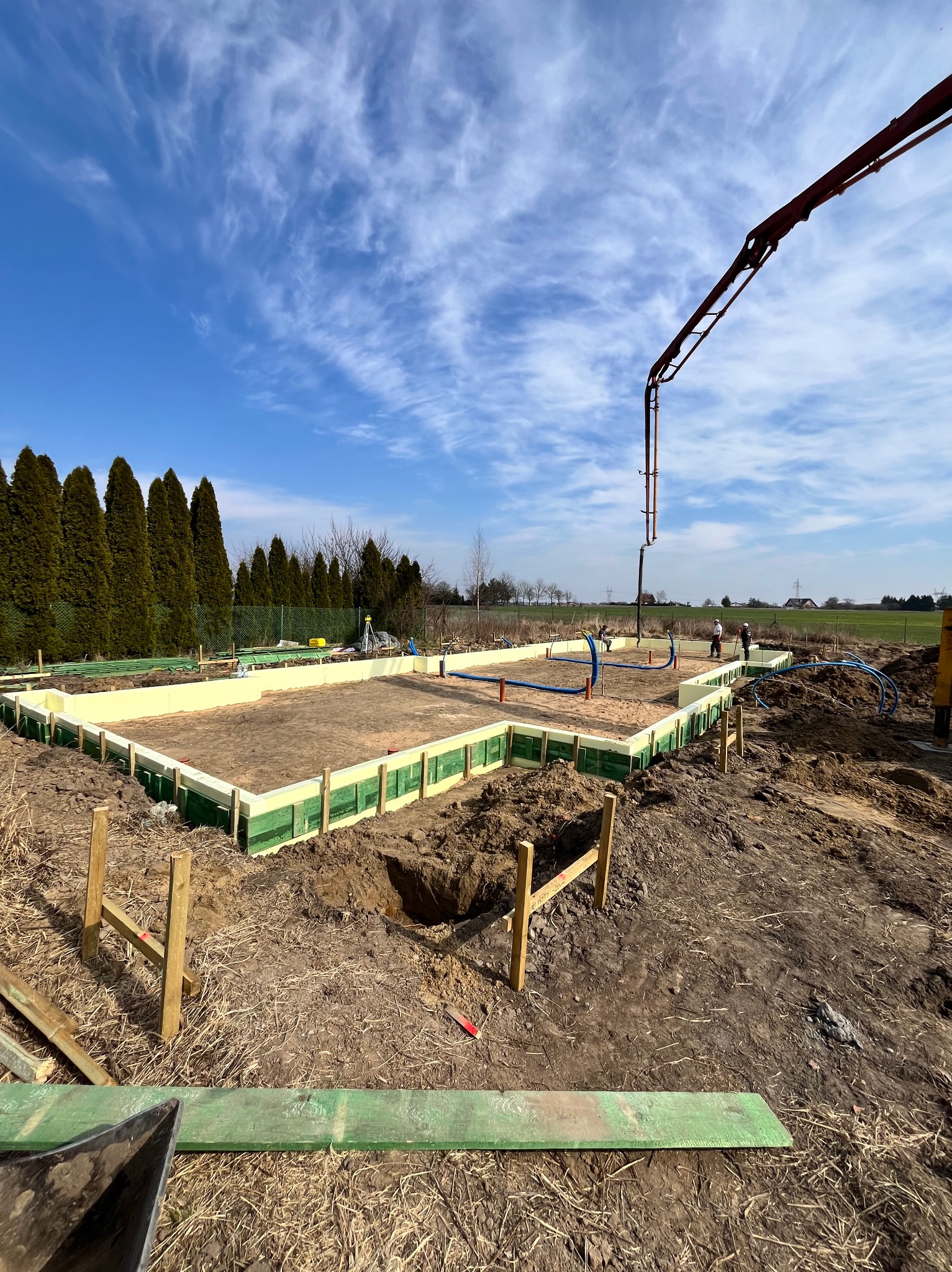 Przygotowany fundament z szalunkiem i izolacją termiczną, w tle ramię pompy do betonu. Widoczne prace przygotowawcze na placu budowy w słoneczny dzień.