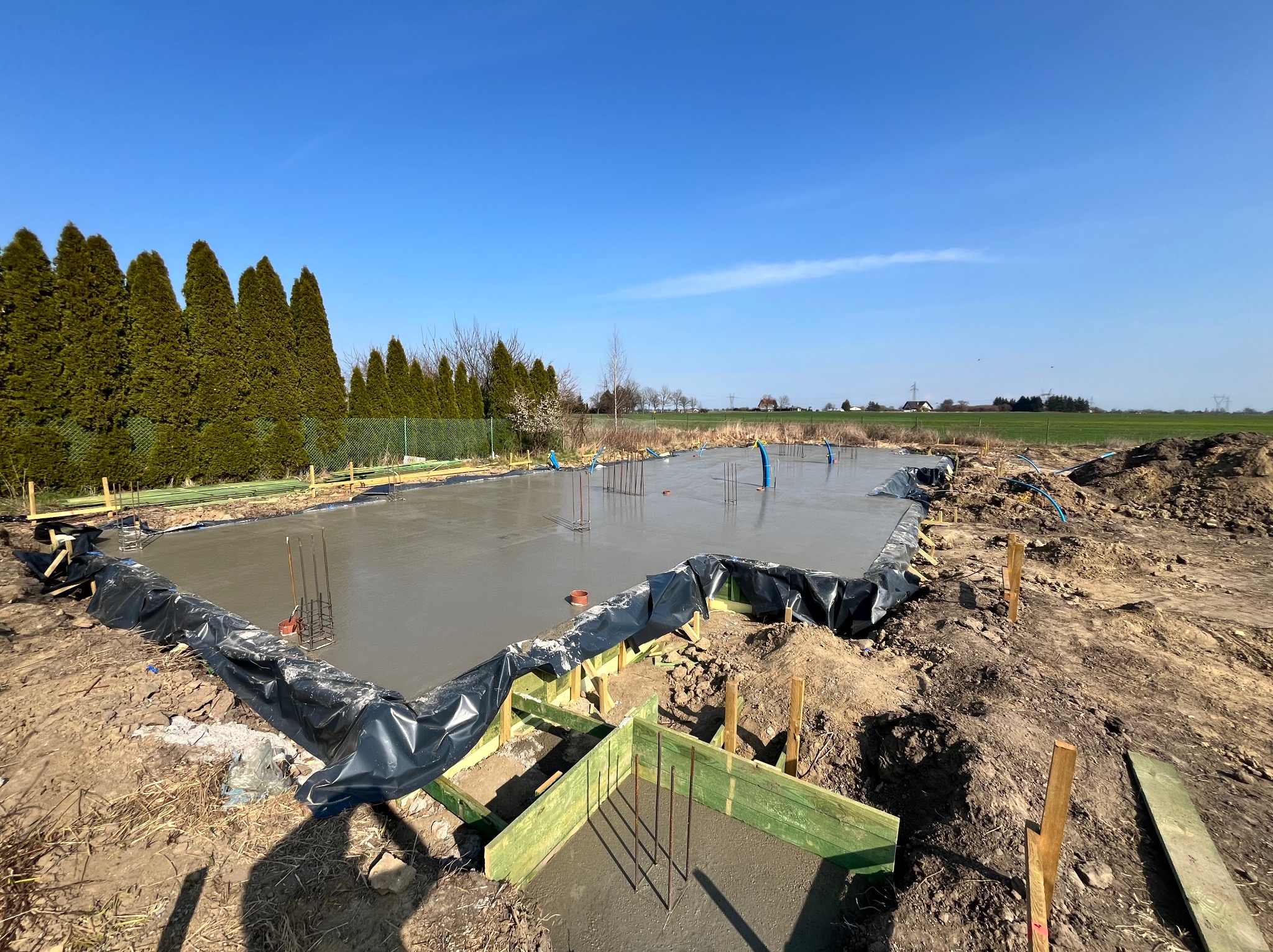 Wylany fundament z wystającymi prętami zbrojeniowymi, zabezpieczony czarną folią, otoczony drewnianym szalunkiem. W tle rząd drzew i błękitne niebo.