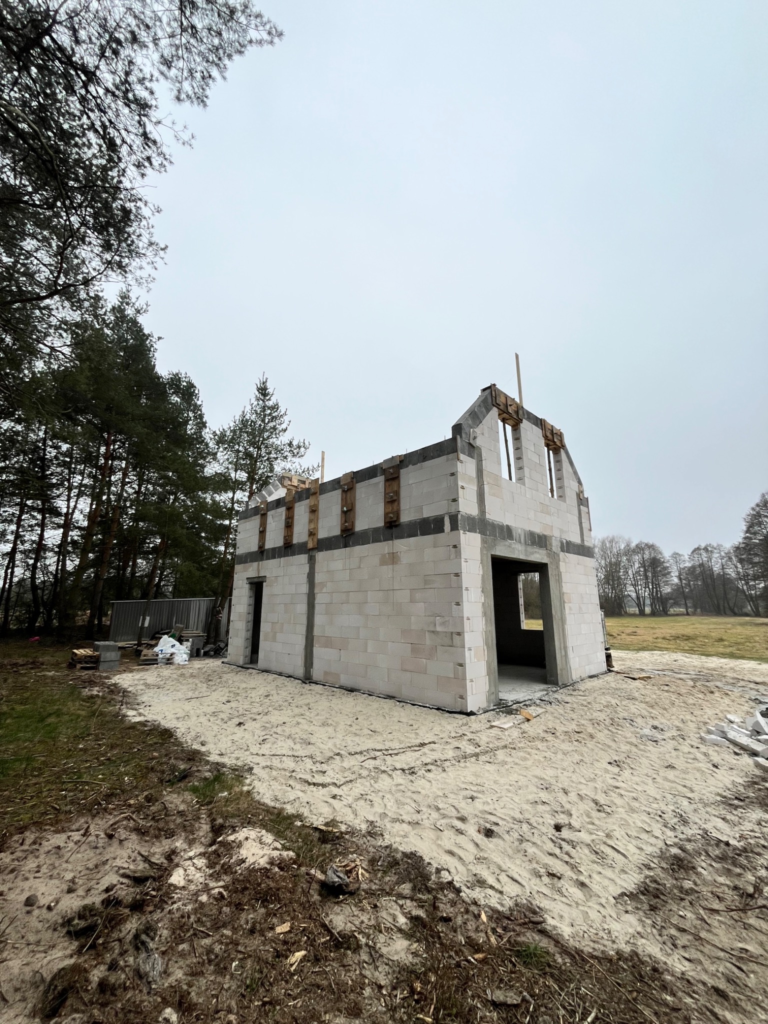 Surowy stan domu murowanego z betonu komórkowego, widok z zewnątrz na tle drzew i nieba. Widoczne otwory okienne i drzwiowe, teren piaszczysty.