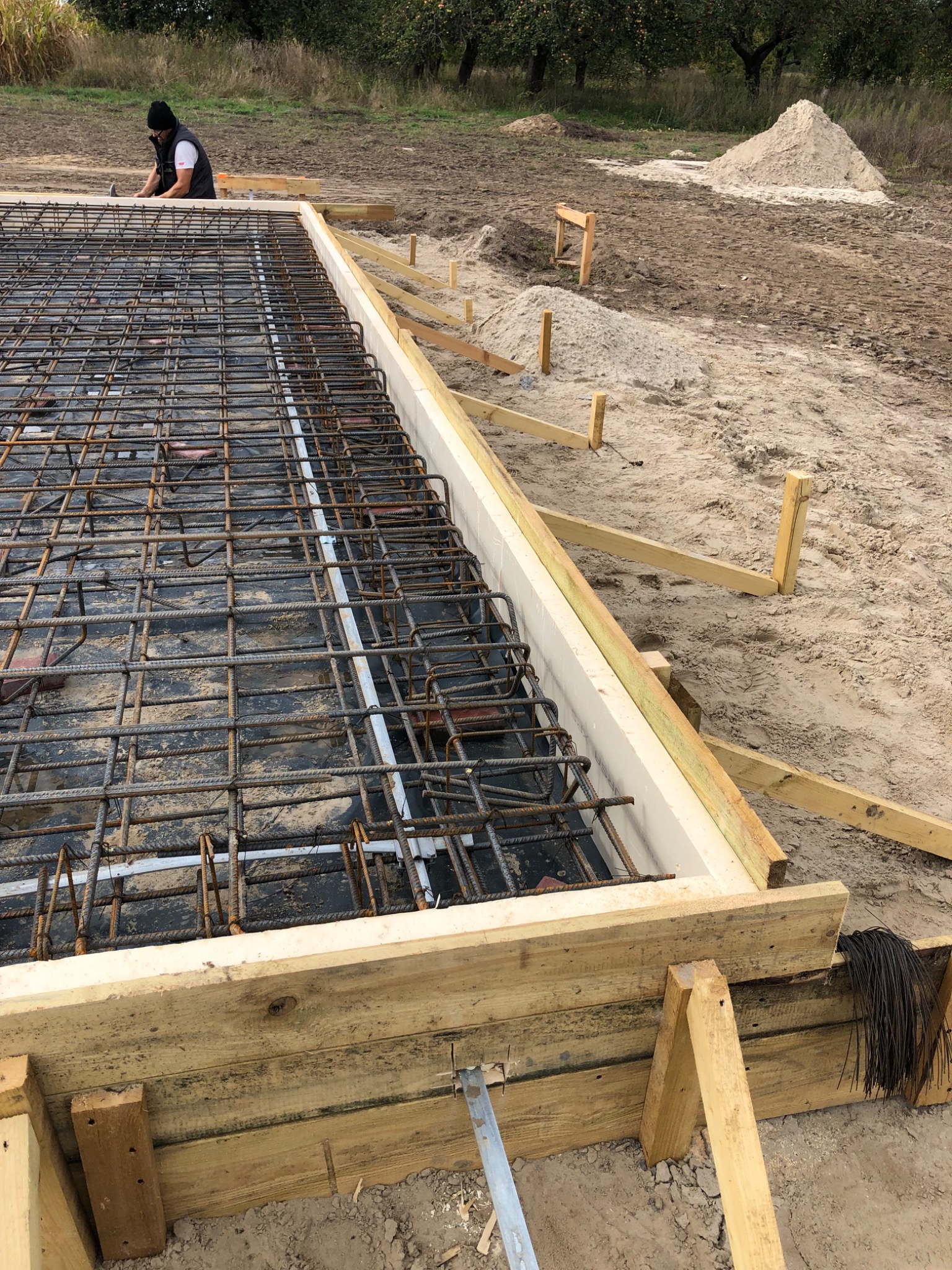 Zbrojenie fundamentu w drewnianym szalunku, widoczny pracownik w oddali, przygotowany teren budowy z pryzmą piasku.