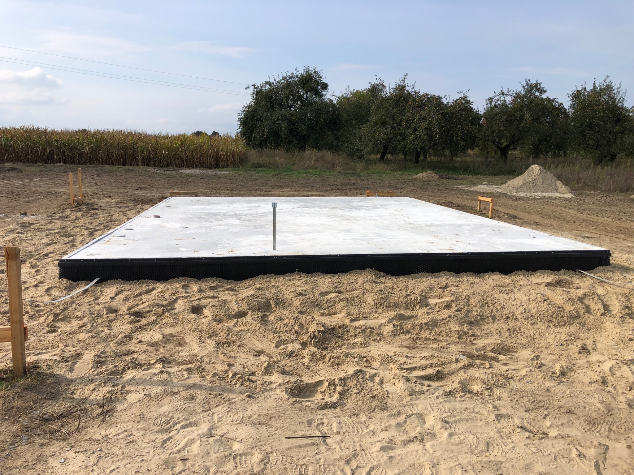 Płyta fundamentowa z czarnym obrzeżem, przygotowana na piaszczystym gruncie, widoczne drewniane kołki wyznaczające obrys, w tle pole kukurydzy i drzewa owocowe.