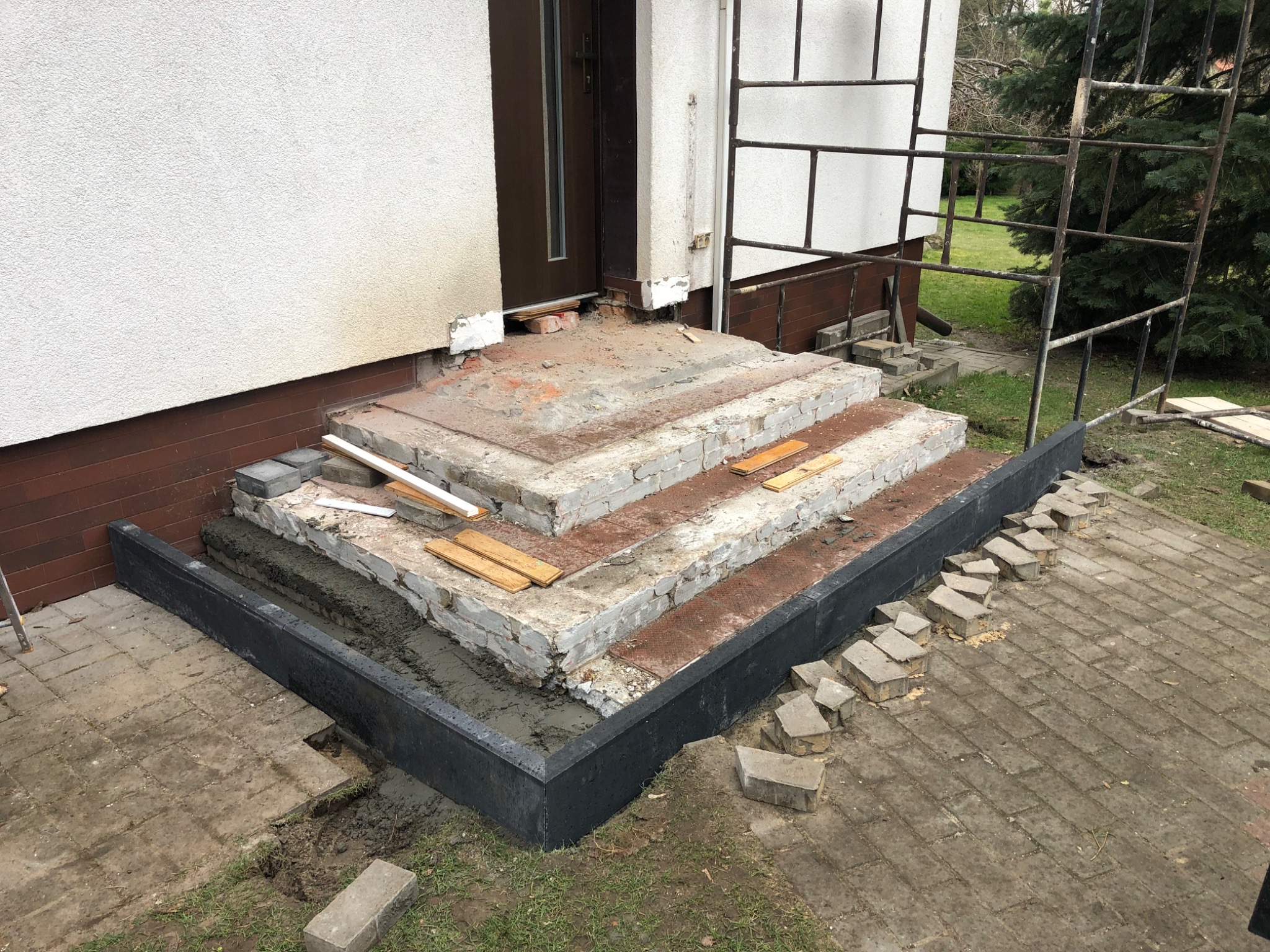 Budowa schodów wejściowych do domu z wykorzystaniem cegieł i zaprawy, widoczne szalunki i częściowo ułożona kostka brukowa przed domem.