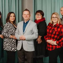 Zespół agencji Social Media Fly pozuje do zdjęcia na tle turkusowej zasłony. Widocznych jest sześć osób, ubrane w różnorodne stroje, od eleganckich marynarek po wzorzyste bluzki. Wszyscy uśmiechają...