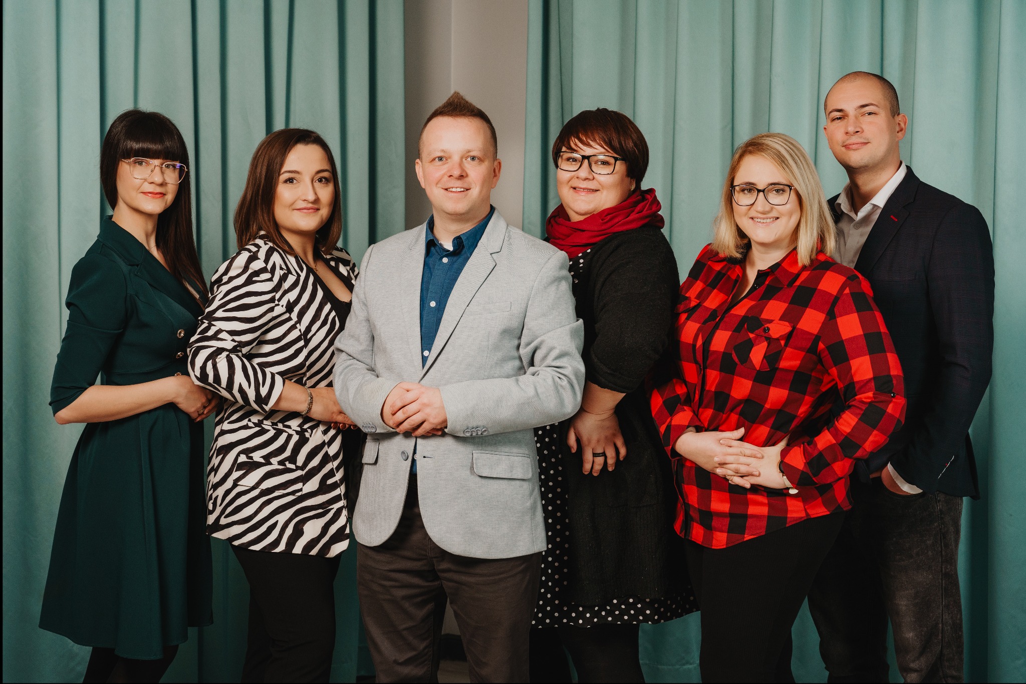 Zespół agencji Social Media Fly pozuje do zdjęcia na tle turkusowej zasłony. Widocznych jest sześć osób, ubrane w różnorodne stroje, od eleganckich marynarek po wzorzyste bluzki. Wszyscy uśmiechają...