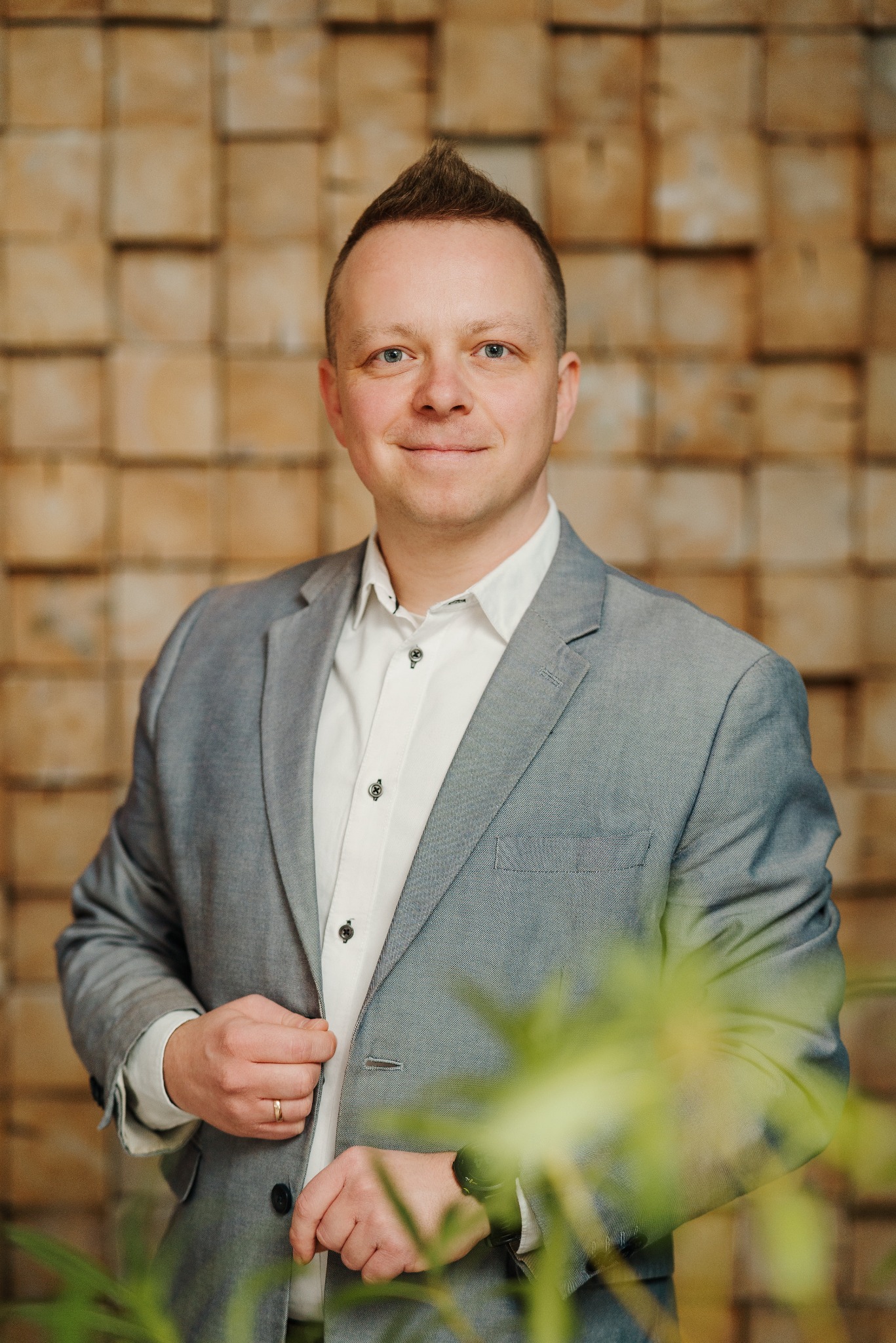 Portret mężczyzny w szarej marynarce i białej koszuli na tle drewnianej ściany w Tczewie, uśmiechający się do kamery, z delikatnie widocznymi roślinami w dolnej części kadru.