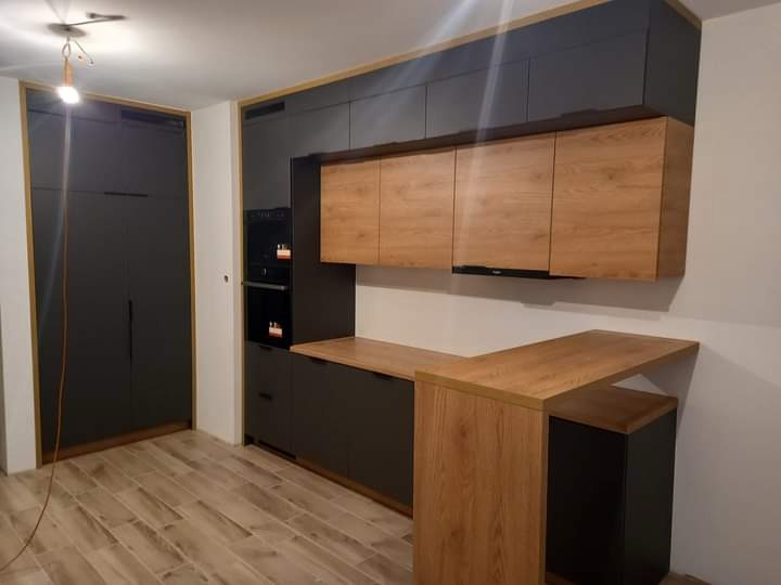 Nowoczesna kuchnia z ciemnoszarymi szafkami dolnymi i górnymi, akcentowana drewnianymi frontami szafek wiszących i blatem, z widocznym oświetleniem podszafkowym i płytkami podłogowymi imitującymi...