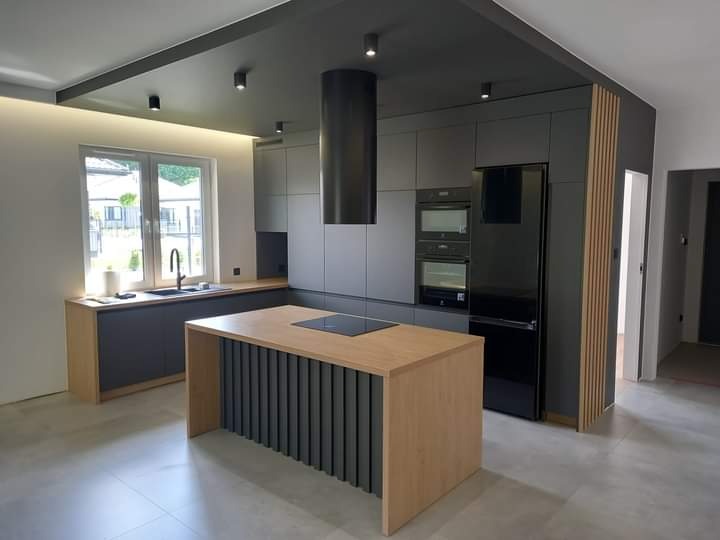 Nowoczesna kuchnia z szarymi szafkami, drewnianym blatem i wyspą, czarnym okapem cylindrycznym i oświetleniem punktowym na ciemnym suficie.