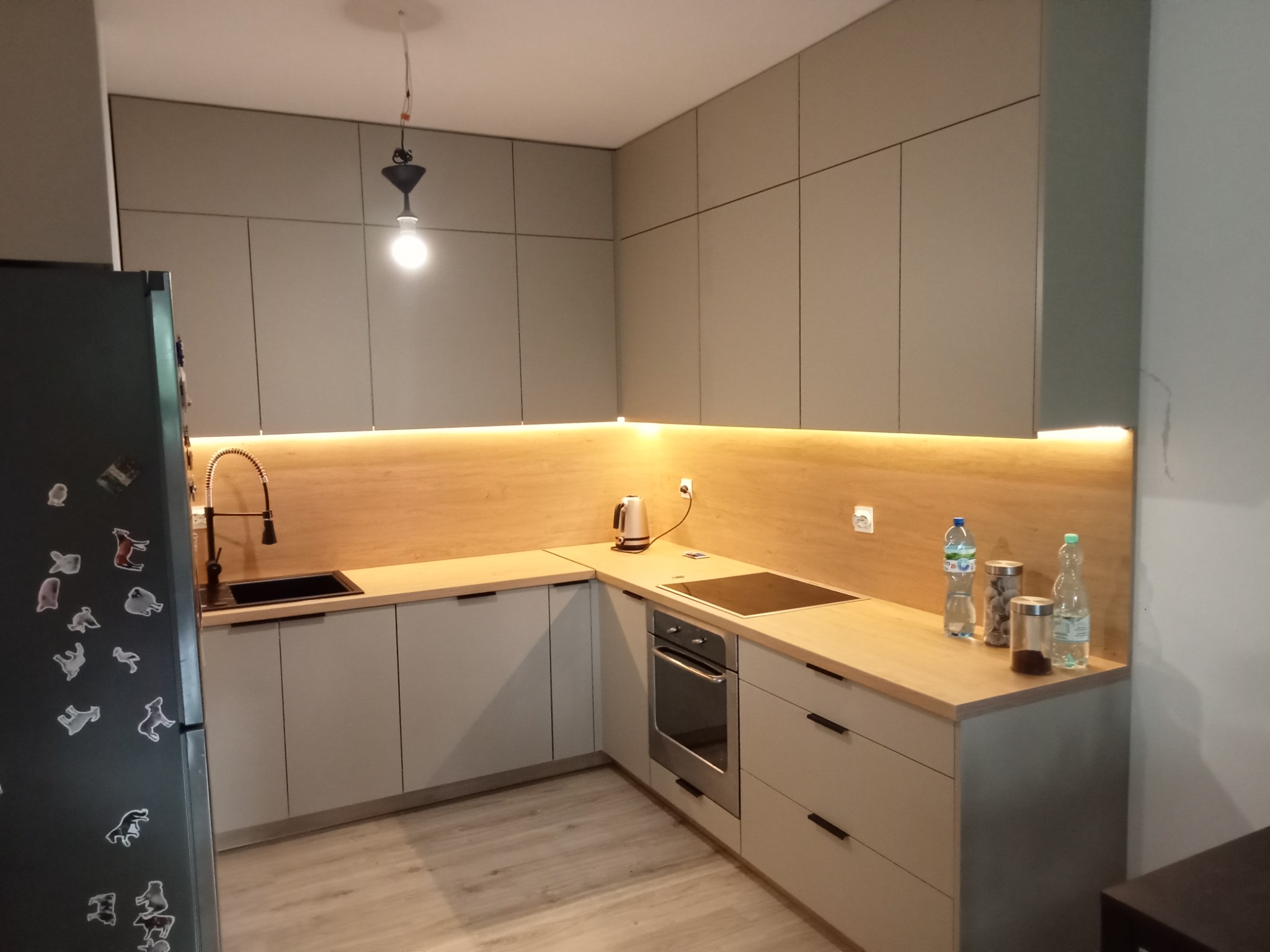 Nowoczesna kuchnia z szarymi szafkami, drewnianym blatem i podświetleniem LED, czarny zlew i bateria, lodówka z magnesami, widok na płytę indukcyjną i piekarnik.
