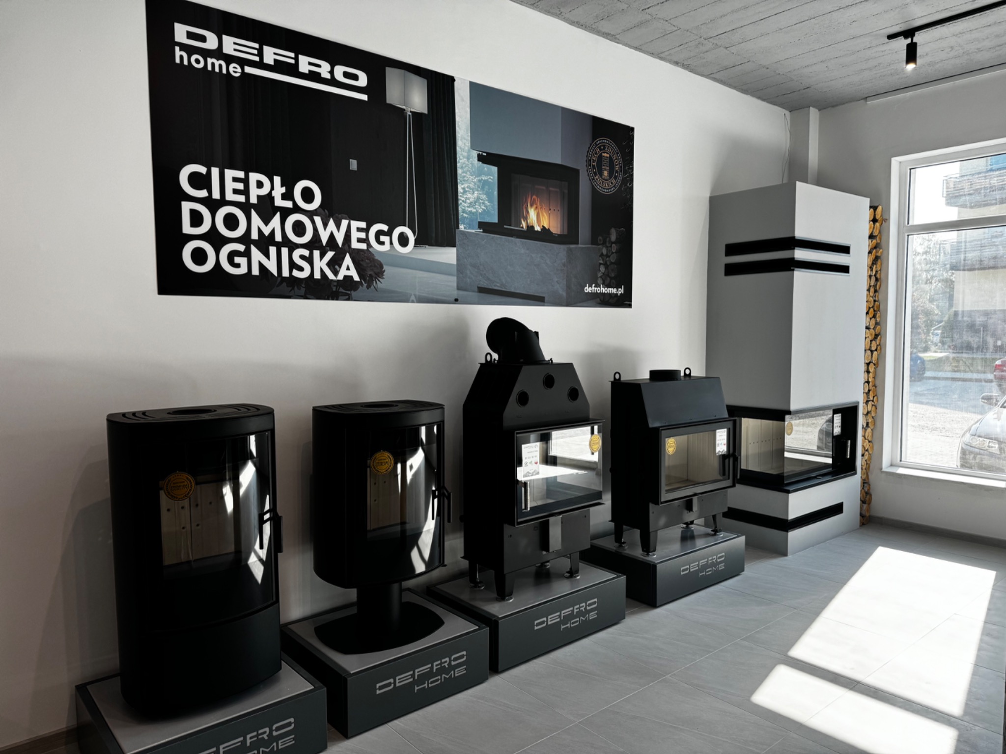 Ekspozycja czterech różnych modeli pieców wolnostojących marki Defro Home w salonie sprzedaży, z plakatem reklamowym w tle i dekoracyjnym kominkiem z drewnem przy oknie.