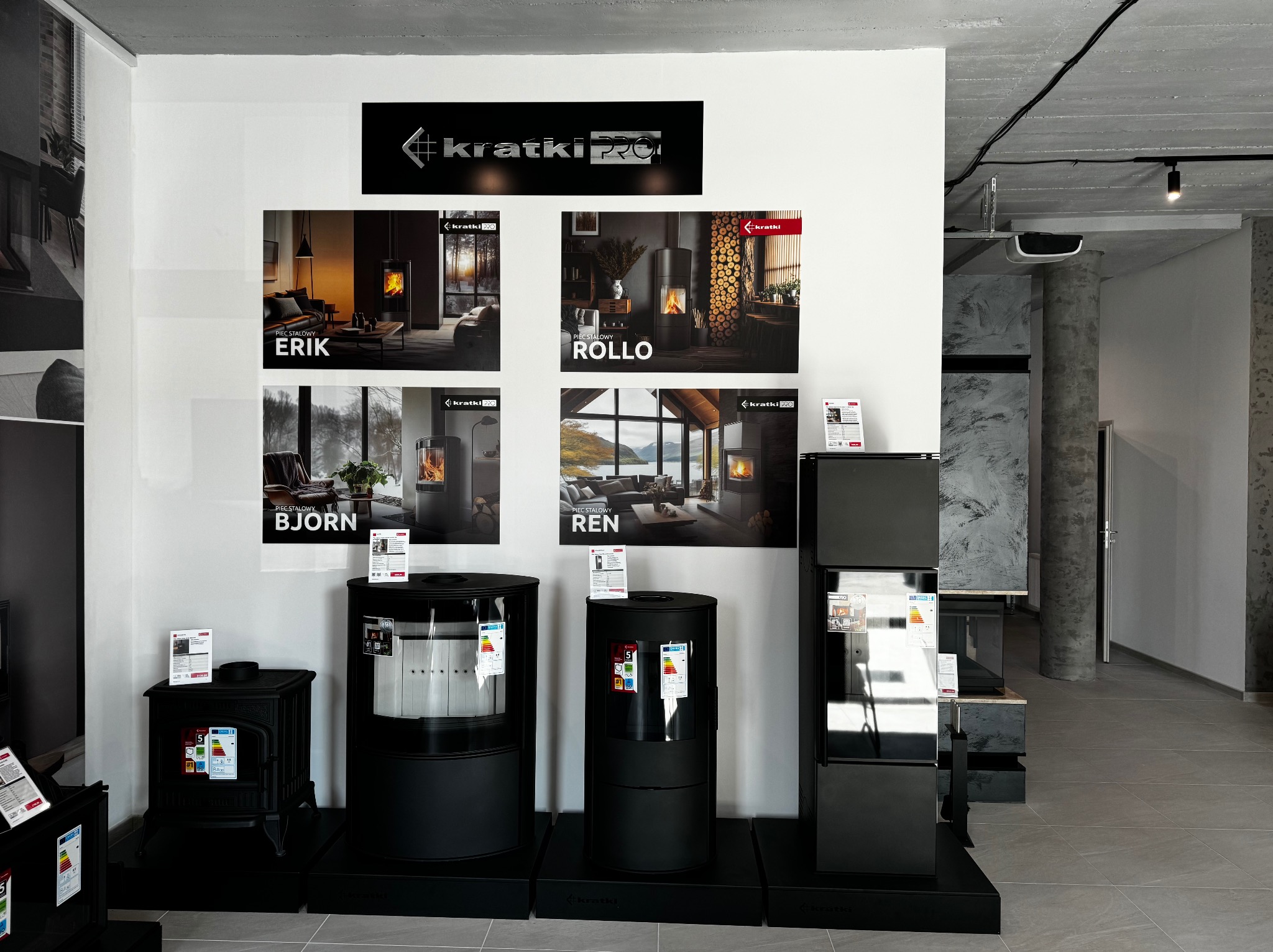 Ekspozycja w salonie: cztery modele pieców stalowych marki Kratki, każdy z plakatem prezentującym go w aranżacji wnętrza. Modele: Erik, Rollo, Bjorn, Ren.