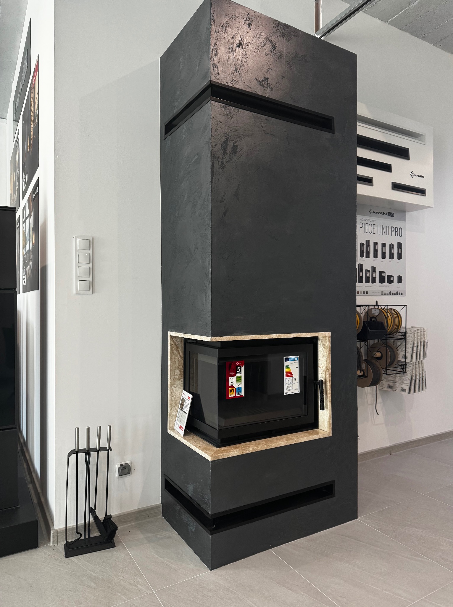 Czarny, nowoczesny piec z obudową imitującą beton, otoczony beżowym kamieniem, z szybą i kompletem akcesoriów do czyszczenia obok.