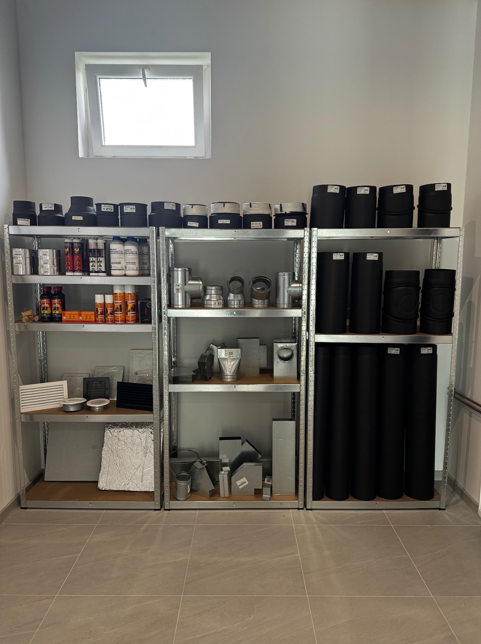 Półki magazynowe z metalowymi elementami instalacyjnymi: rury, kolanka, trójniki, kształtki, redukcje, puszki, preparaty chemiczne i uszczelniacze.
