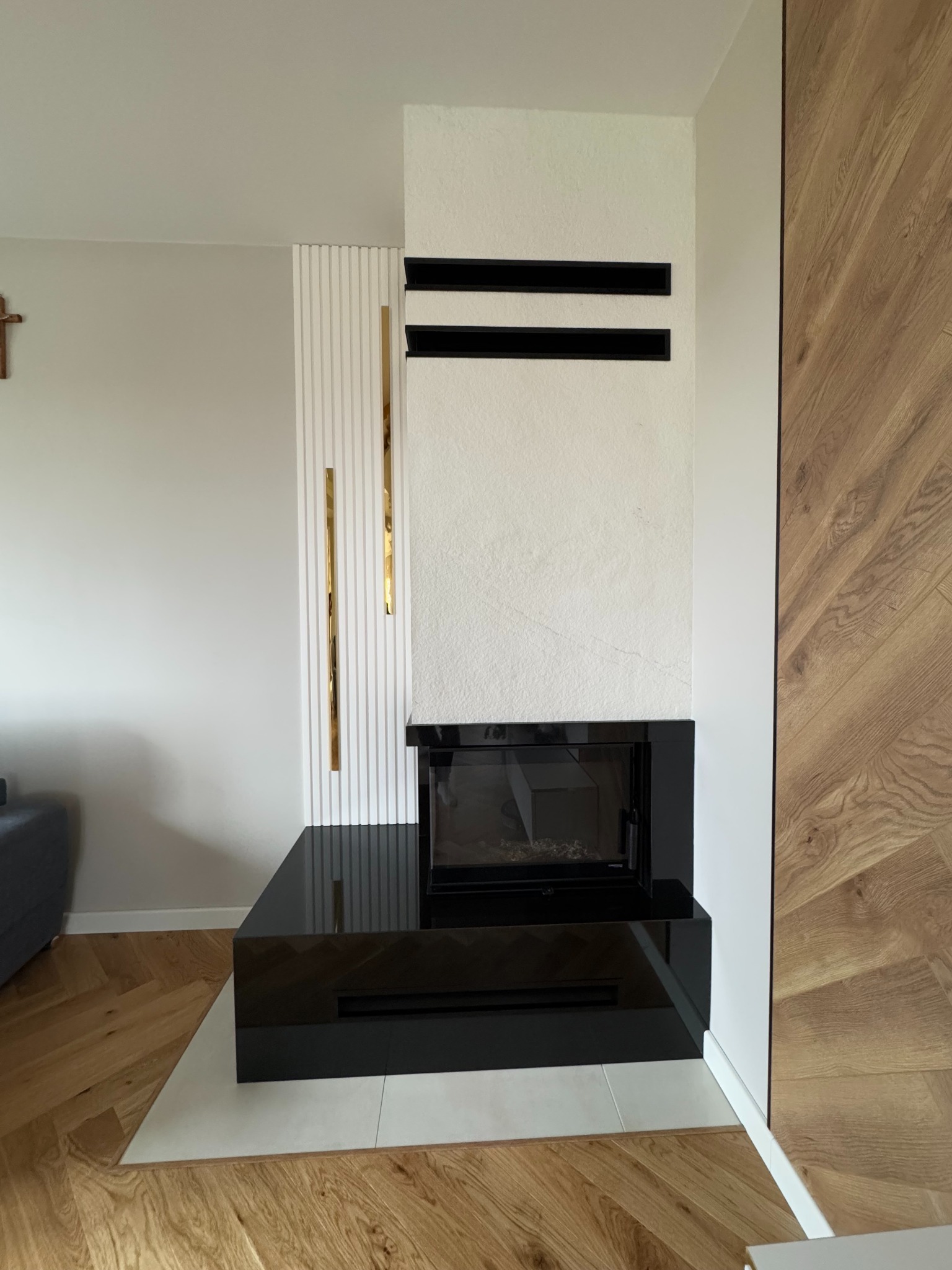 Nowoczesny wkład kominkowy DefroHome obudowany czarnym granitem, z elementami dekoracyjnymi w kolorze złota, na tle parkietu ułożonego w jodełkę.