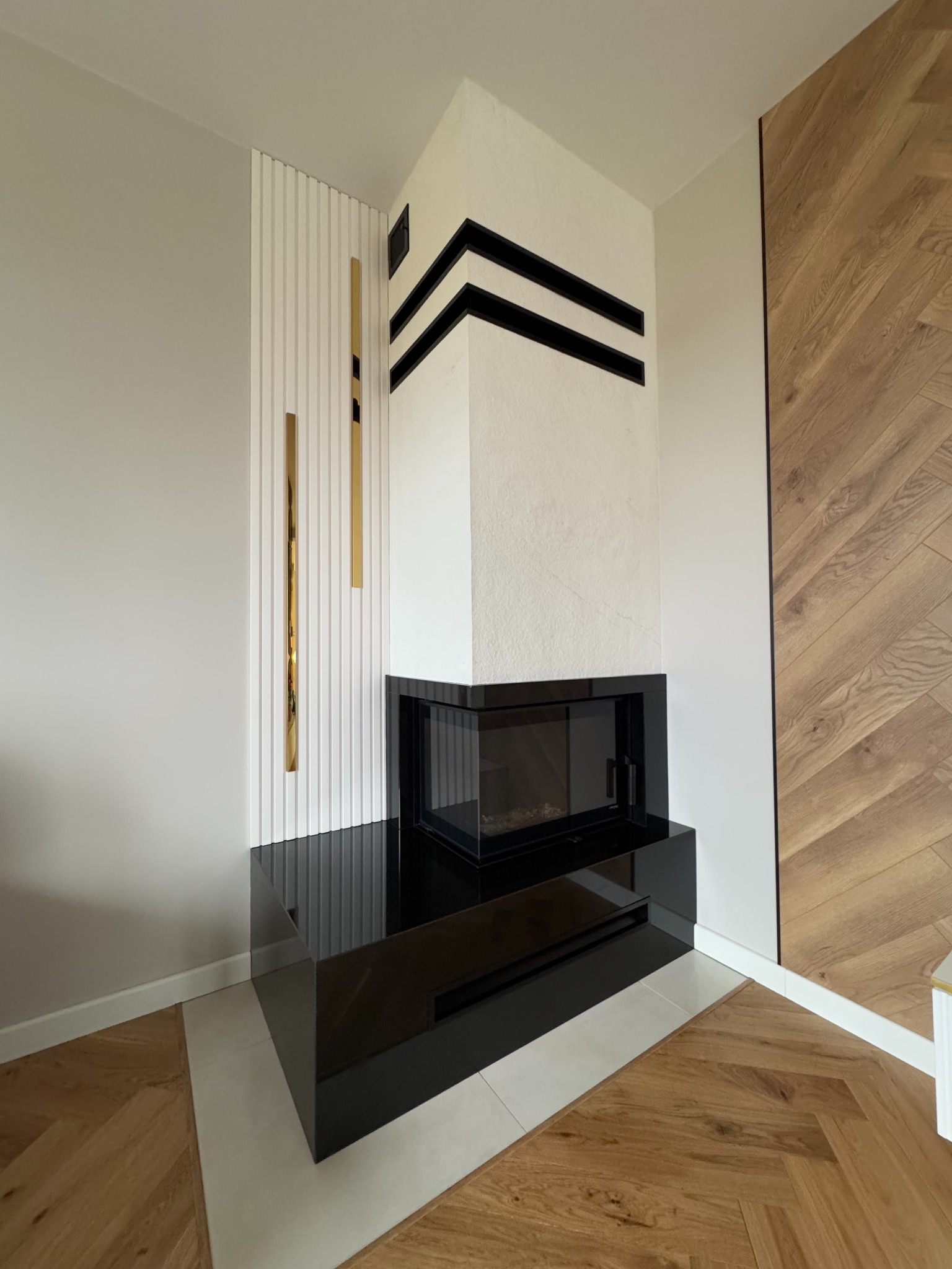 Nowoczesny biokominek narożny z czarną obudową i białym korpusem, dekorowany poziomymi czarnymi pasami, umieszczony w pomieszczeniu z parkietem ułożonym w jodełkę.