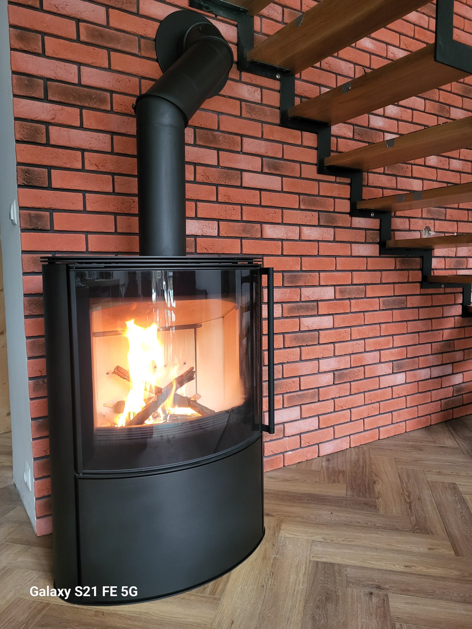 Nowoczesny, czarny piec wolnostojący z palącym się drewnem, ustawiony na drewnianej podłodze w jodełkę, z ceglaną ścianą i drewnianymi schodami w tle.