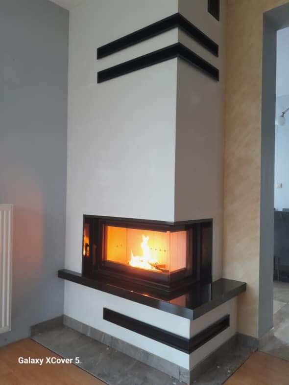 Nowoczesny kominek narożny z czarnymi akcentami i płomieniami, widok z boku, ciepłe światło ognia kontrastuje z chłodnymi odcieniami ścian.