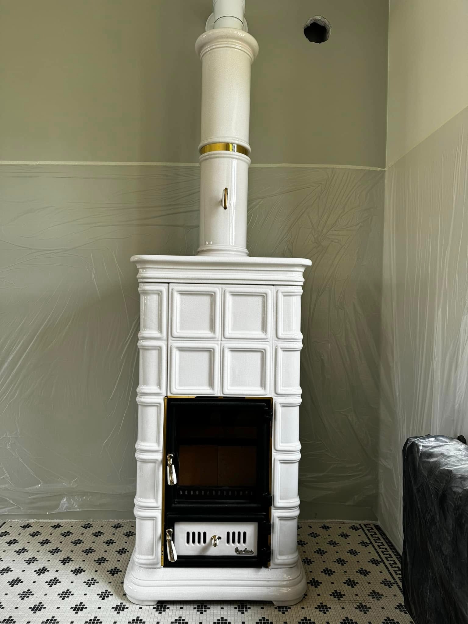 Biały piec kaflowy z czarnym wkładem, widoczny podczas montażu, ściany zabezpieczone folią malarską, na podłodze biało-czarna mozaika.
