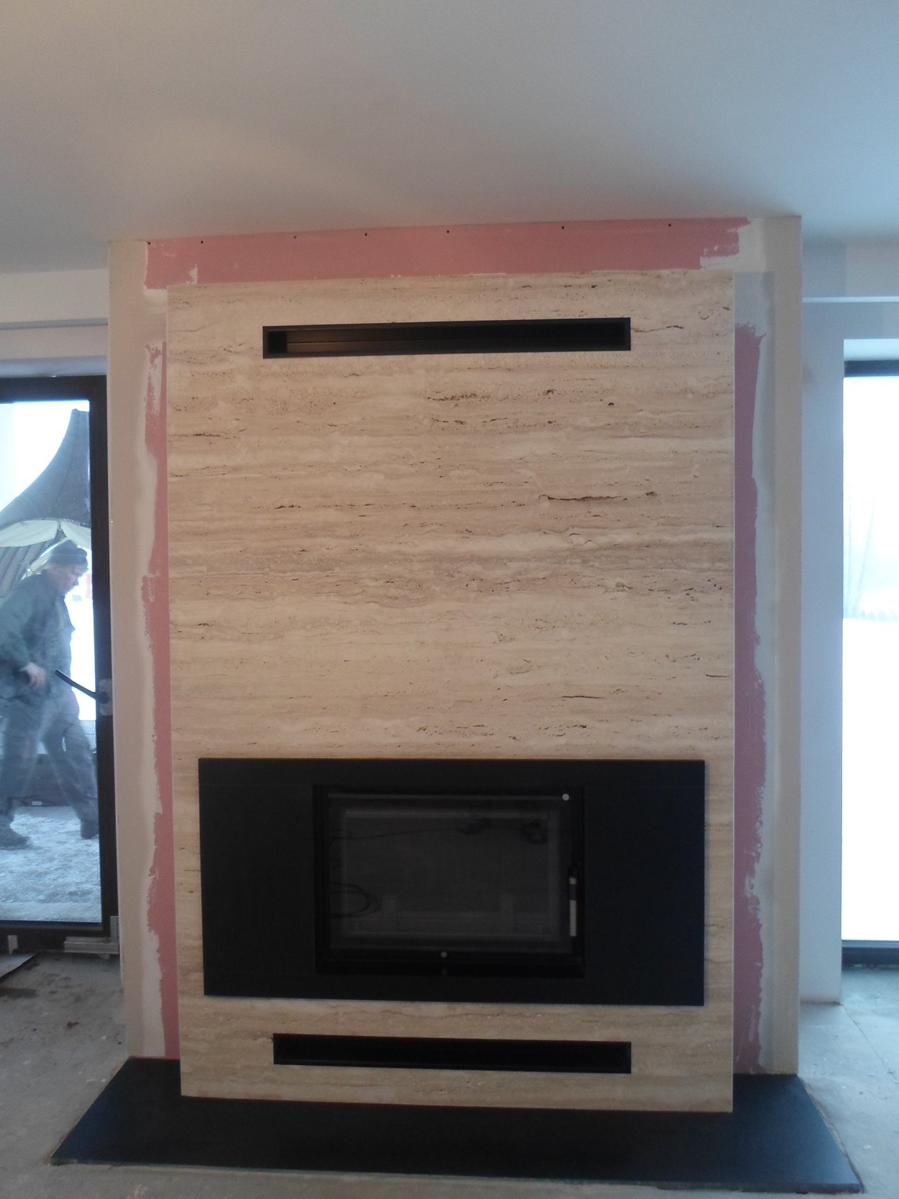 Montaż obudowy z trawertynu wokół wkładu kominkowego z czarną ramą, widoczne surowe wykończenie ścian i pracownik w tle.