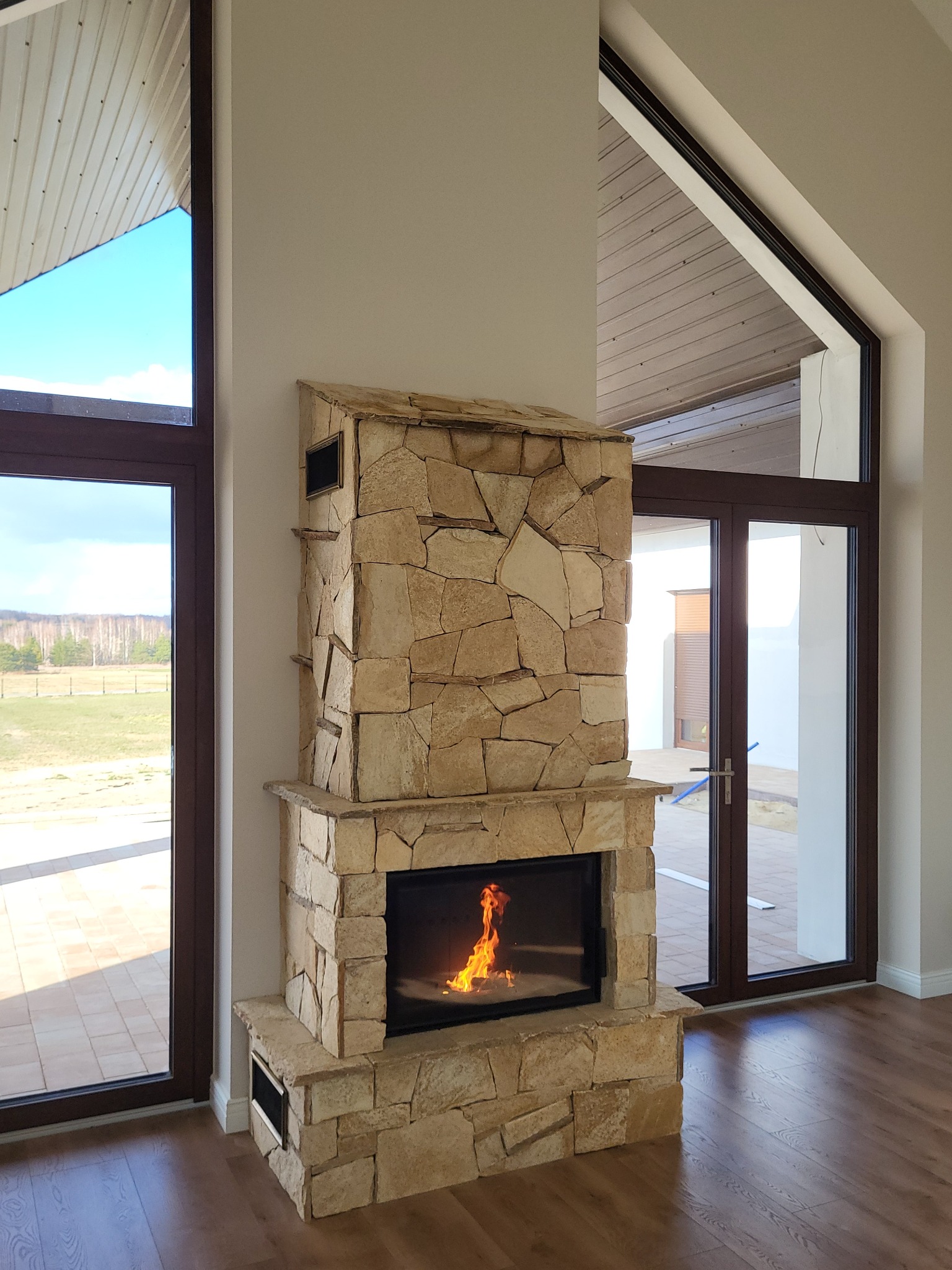 Kamienny piec z paleniskiem w nowoczesnym wnętrzu z dużymi oknami, widoczne płomienie i struktura kamienia.