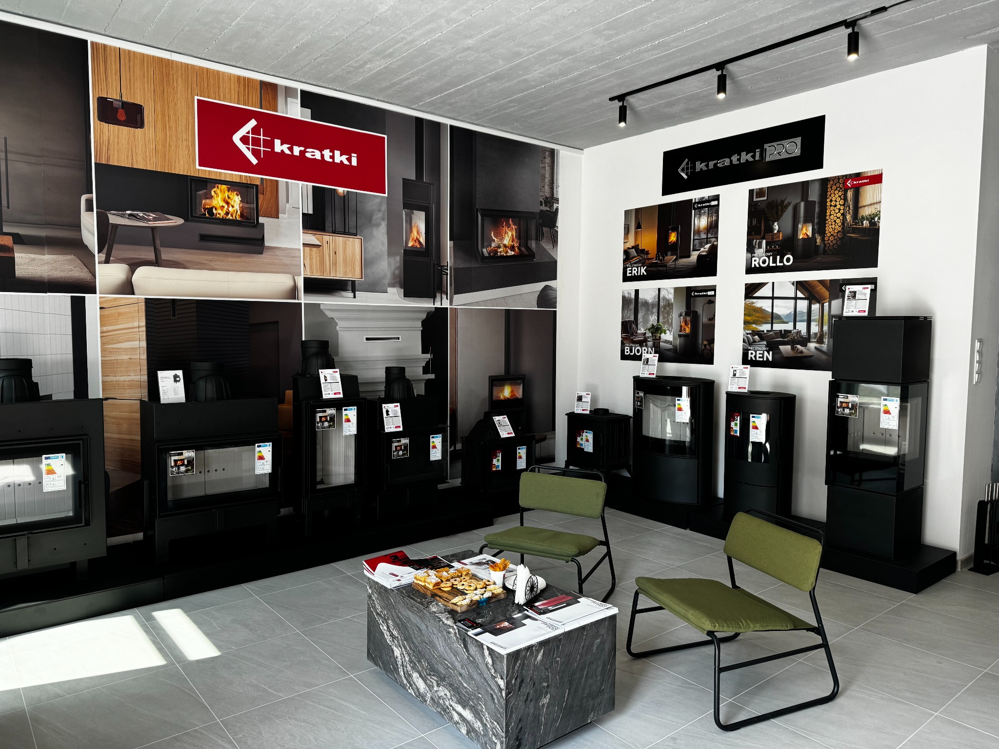 Wnętrze salonu z ekspozycją urządzeń grzewczych marki Kratki, prezentacja modeli z szybą, plakaty reklamowe na ścianie, stolik kawowy z przekąskami i krzesłami dla klientów.