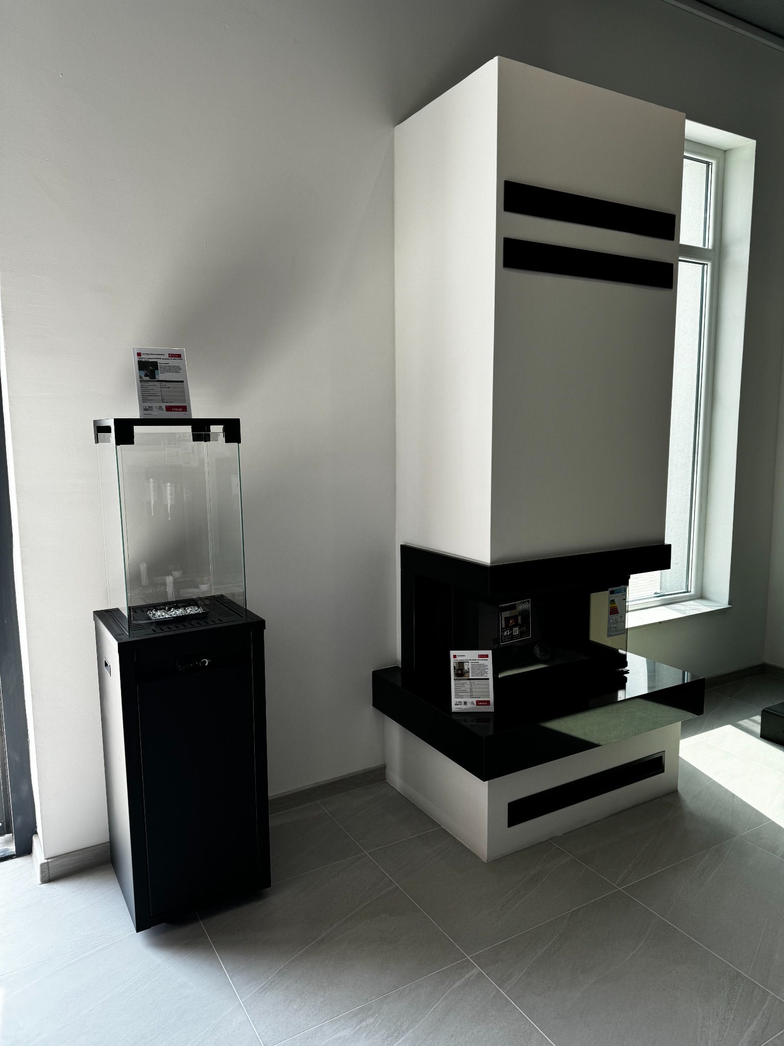 Nowoczesny, minimalistyczny wystrój wnętrza z dwoma modelami biokominków: jeden z przeszkloną obudową na czarnej podstawie, drugi biało-czarny, przypominający kolumnę, z informacjami o produkcie...