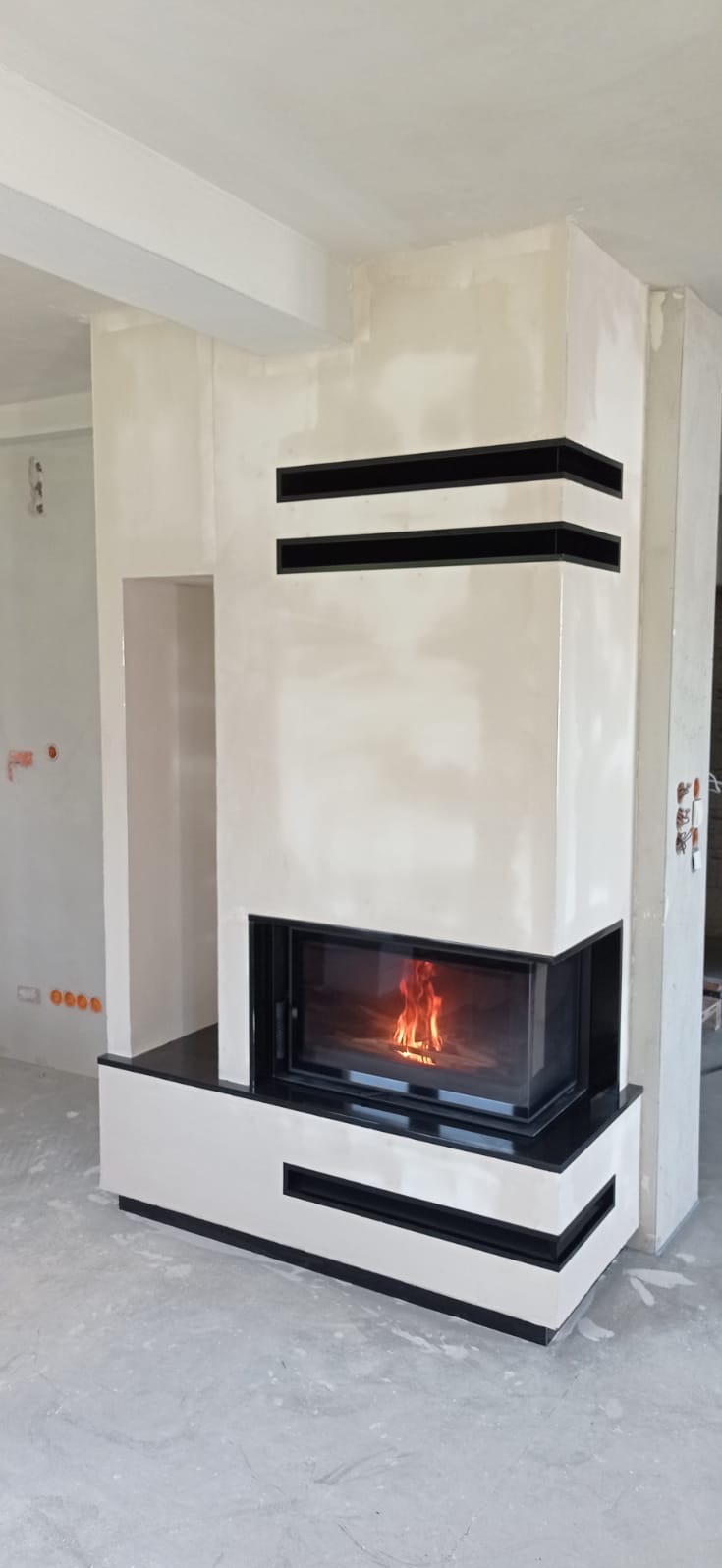 Nowoczesny kominek narożny z czarnymi akcentami i płomieniami, widoczny w surowym wnętrzu, prawdopodobnie w trakcie wykańczania.