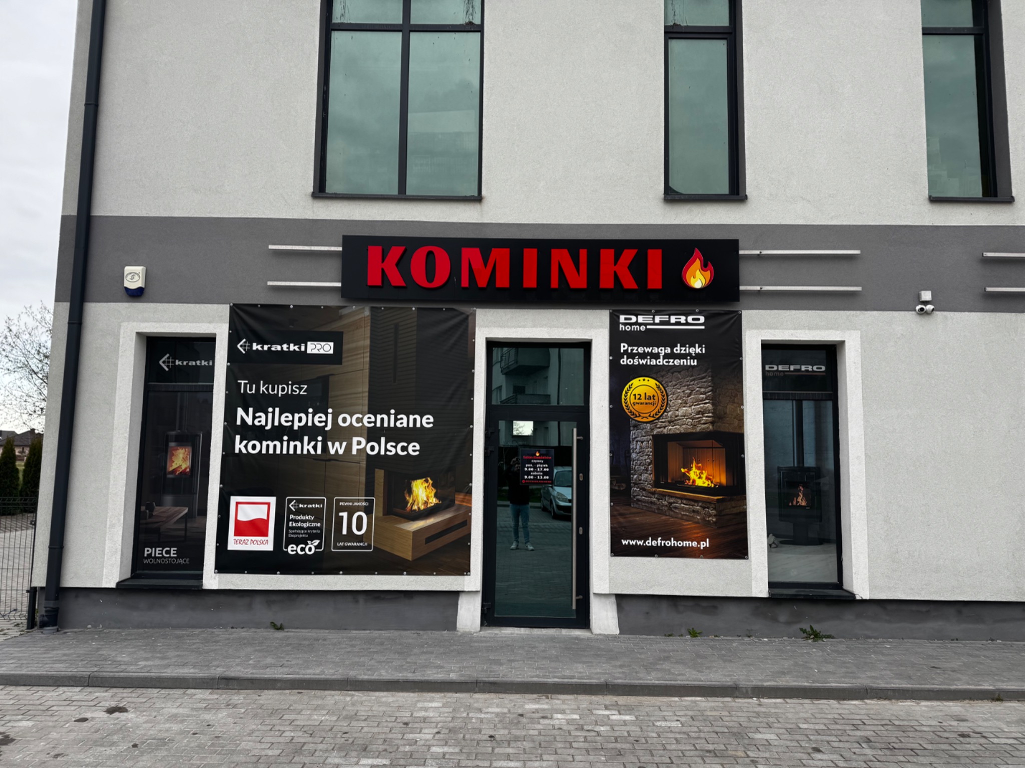 Fasada budynku z szyldem 'KOMINKI' i banerami reklamowymi producentów, prezentującymi modele pieców wolnostojących i wkłady kominkowe. Widoczne logo 'Defro Home'.