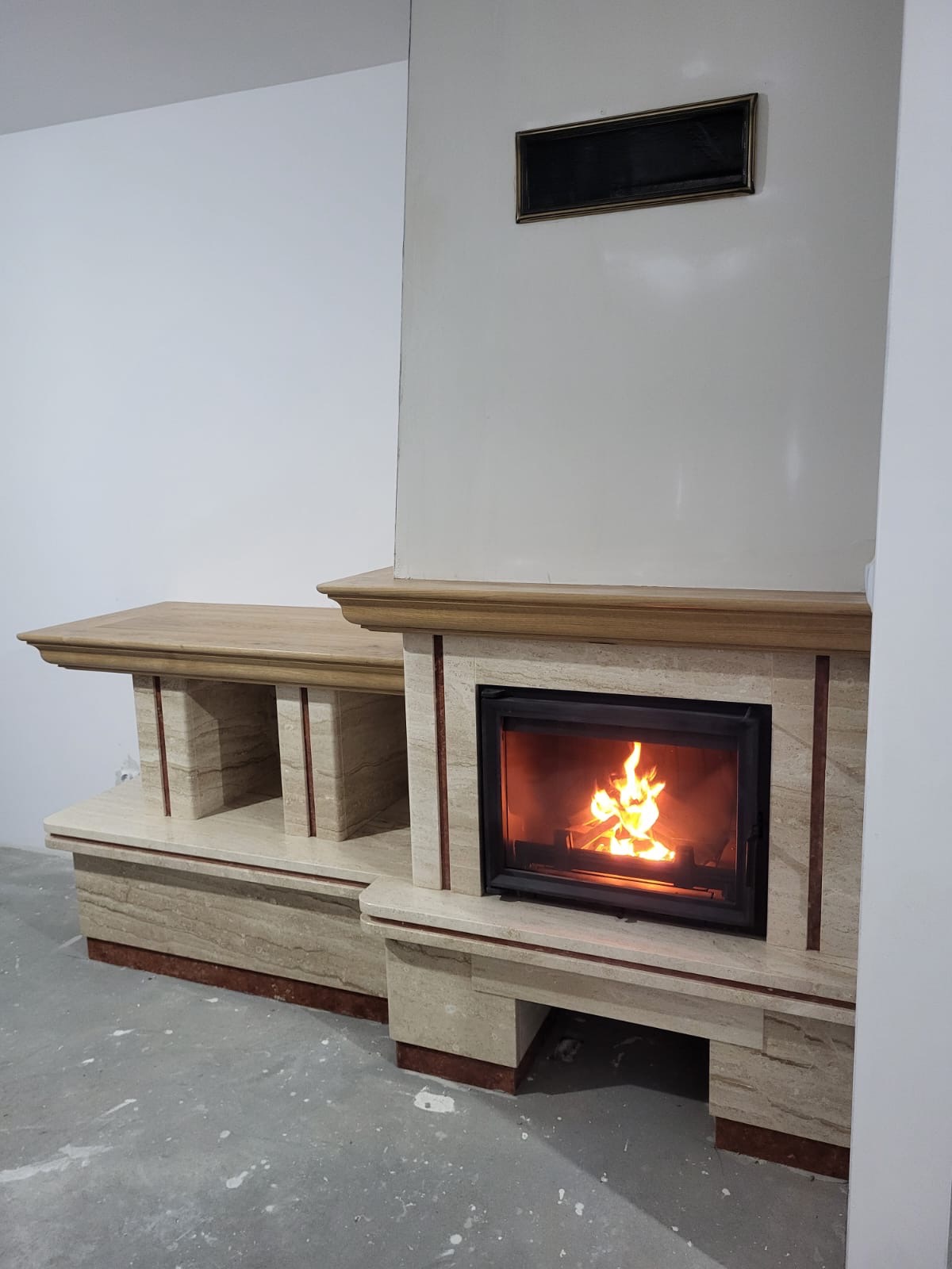 Trawertynowy kominek z paleniskiem i drewnianą półką, ozdobiony bordowymi akcentami, w surowym wnętrzu z szarą podłogą i czarną ramką na ścianie.