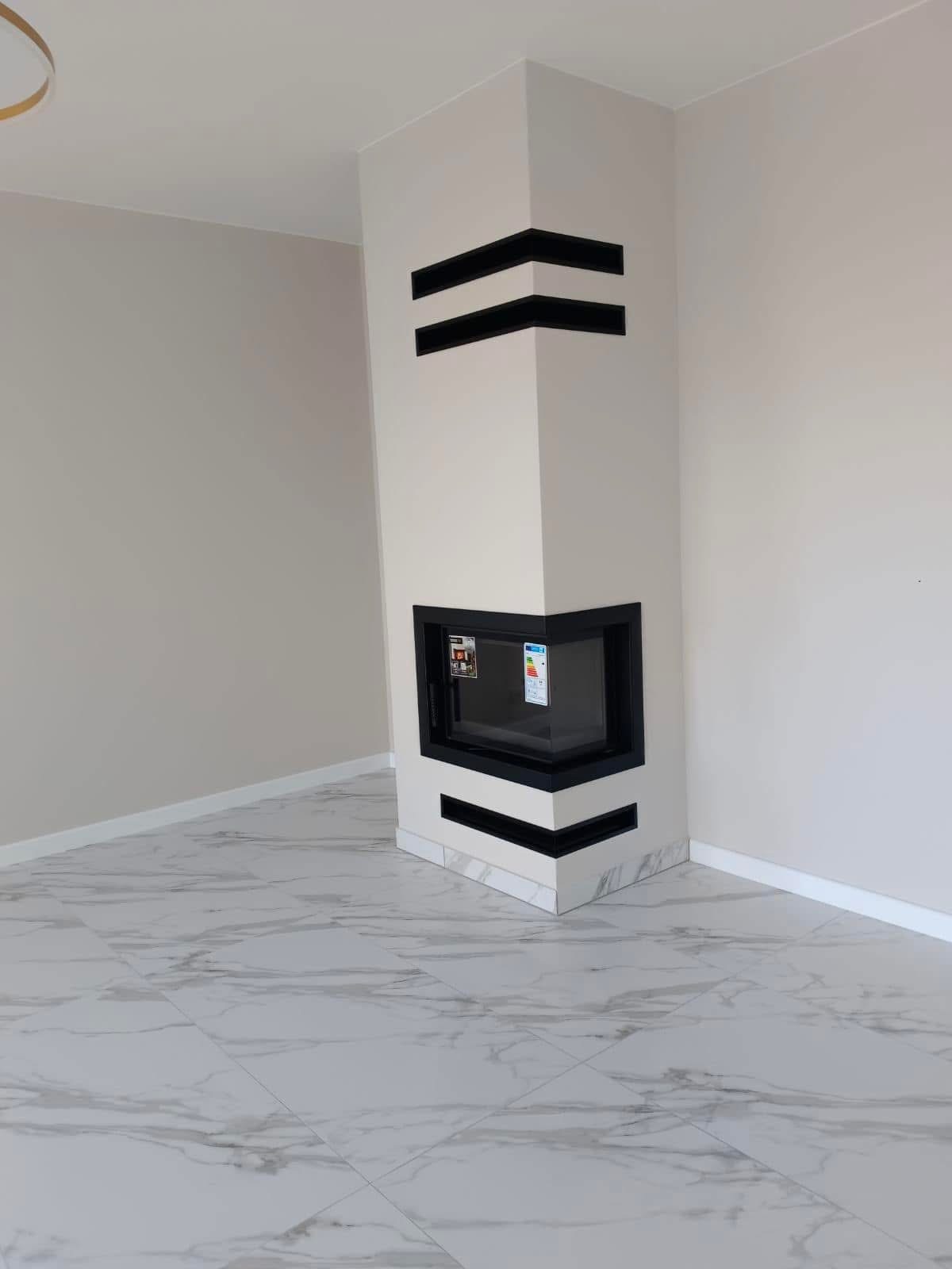 Narożny kominek z czarnymi akcentami w nowoczesnym wnętrzu. Białe ściany i marmurowe płytki podłogowe. Widoczne detale wykończenia.