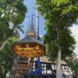 Montaż wieżyczki kościoła za pomocą dźwigu, widoczne drewniane elementy konstrukcyjne i fragment elewacji budynku.