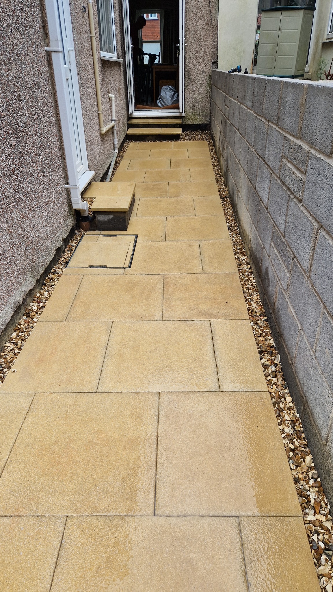 Żółta ścieżka z płyt kamiennych prowadząca do drzwi wejściowych, wykończona drobnymi kamykami po bokach, z widoczną studzienką kanalizacyjną i ścianą z bloczków betonowych.