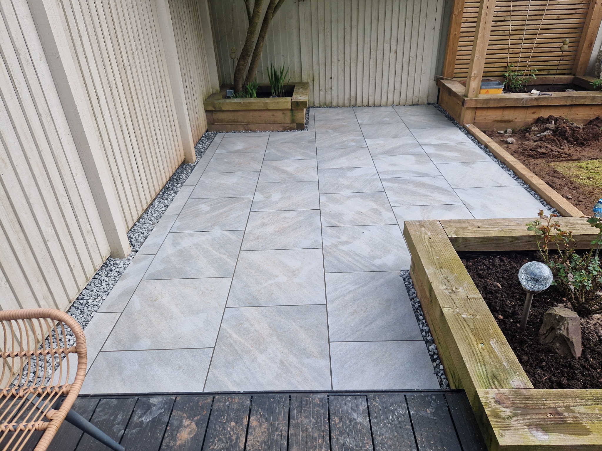 Jasnoszare płyty tarasowe ułożone na zewnątrz, z drewnianymi obrzeżami rabat i kruszywem, widoczny fragment wiklinowego krzesła.