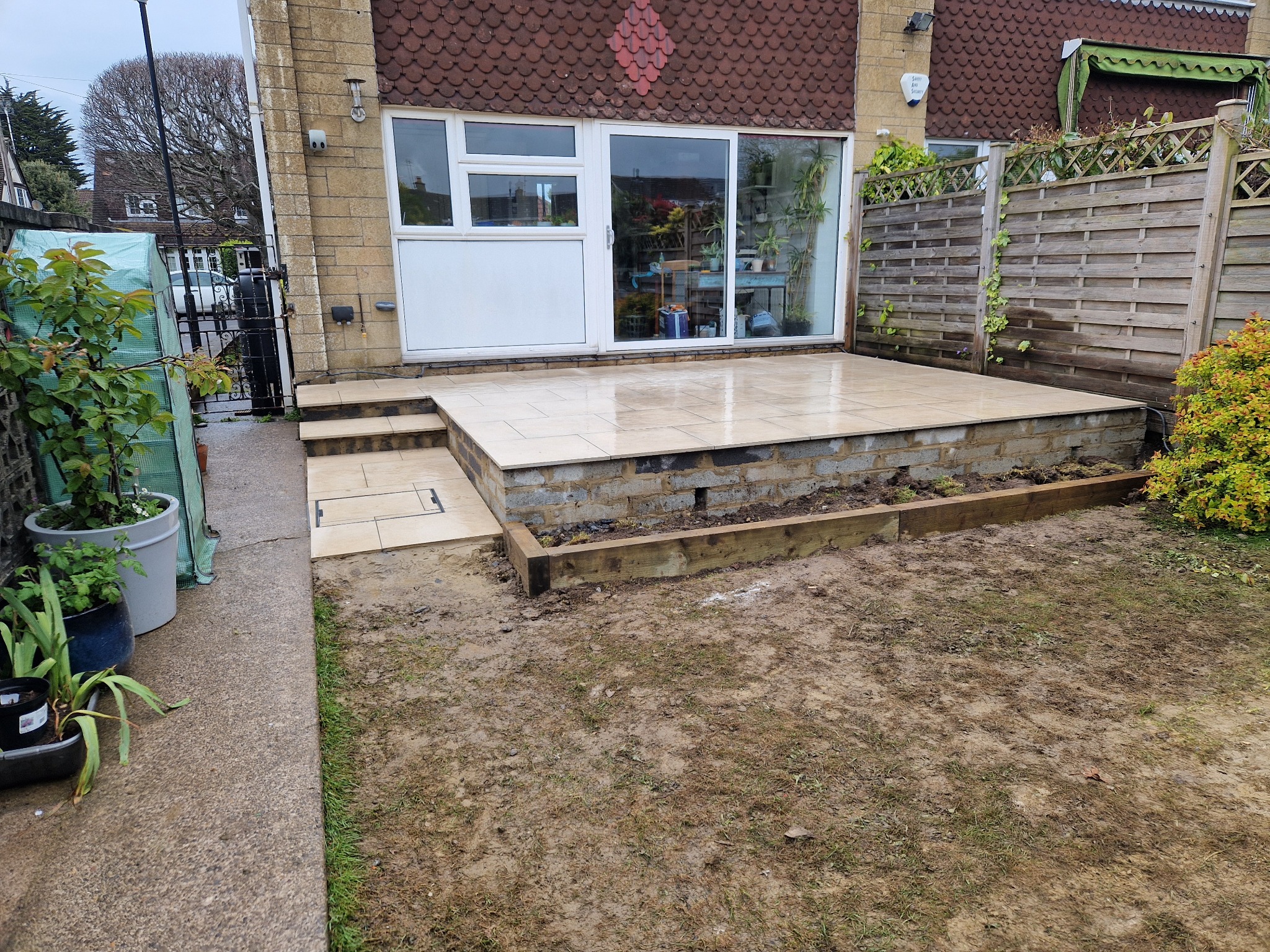 Wyłożony jasnymi płytkami taras z dwoma stopniami, widok na przesuwne drzwi tarasowe i mokry trawnik po deszczu.