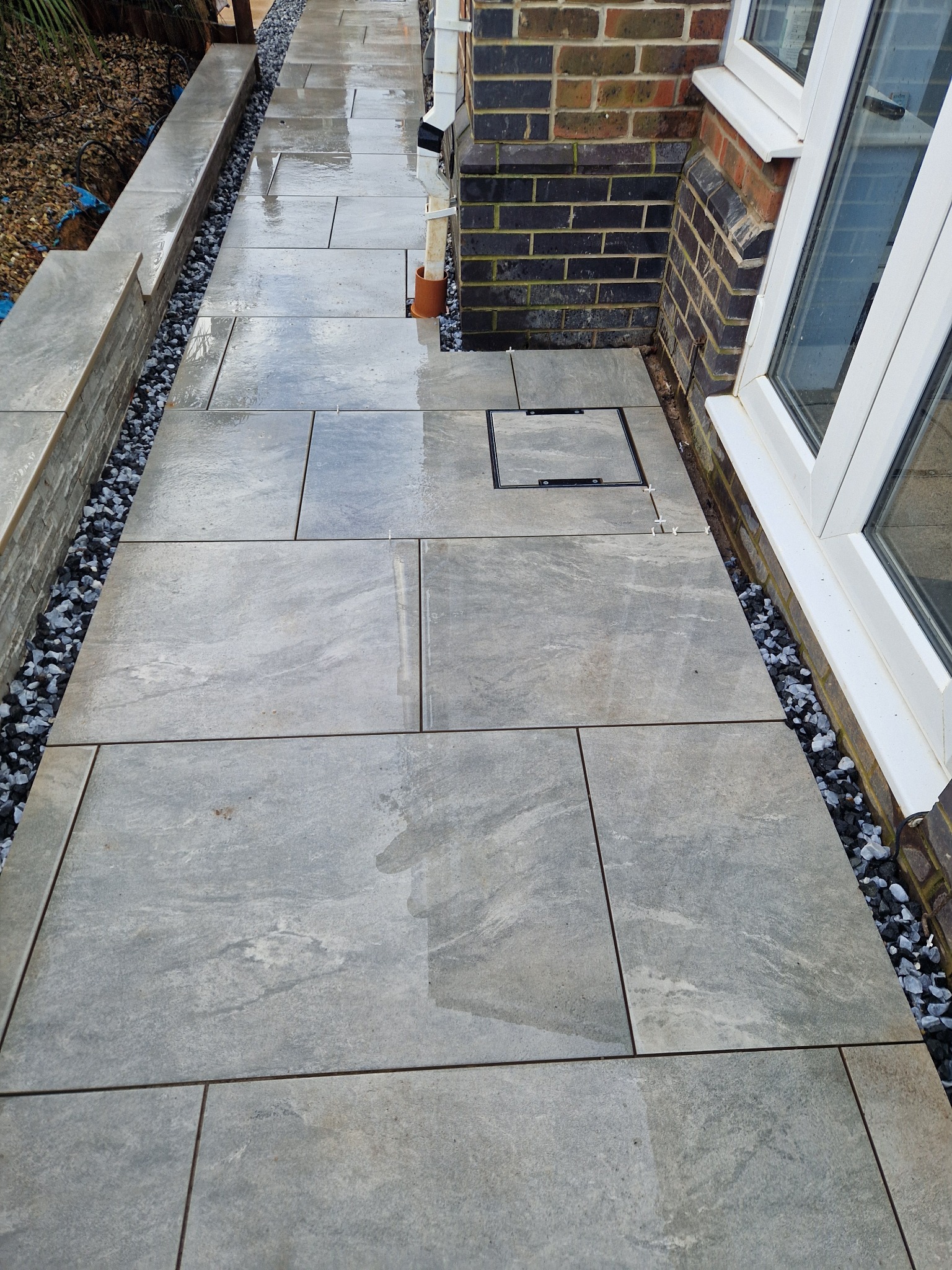 Świeżo ułożony chodnik z szarych, prostokątnych płyt kamiennych, oddzielony od budynku i rabaty pasem drobnych, ciemnych kamyczków. Widoczna studzienka rewizyjna wkomponowana w nawierzchnię.