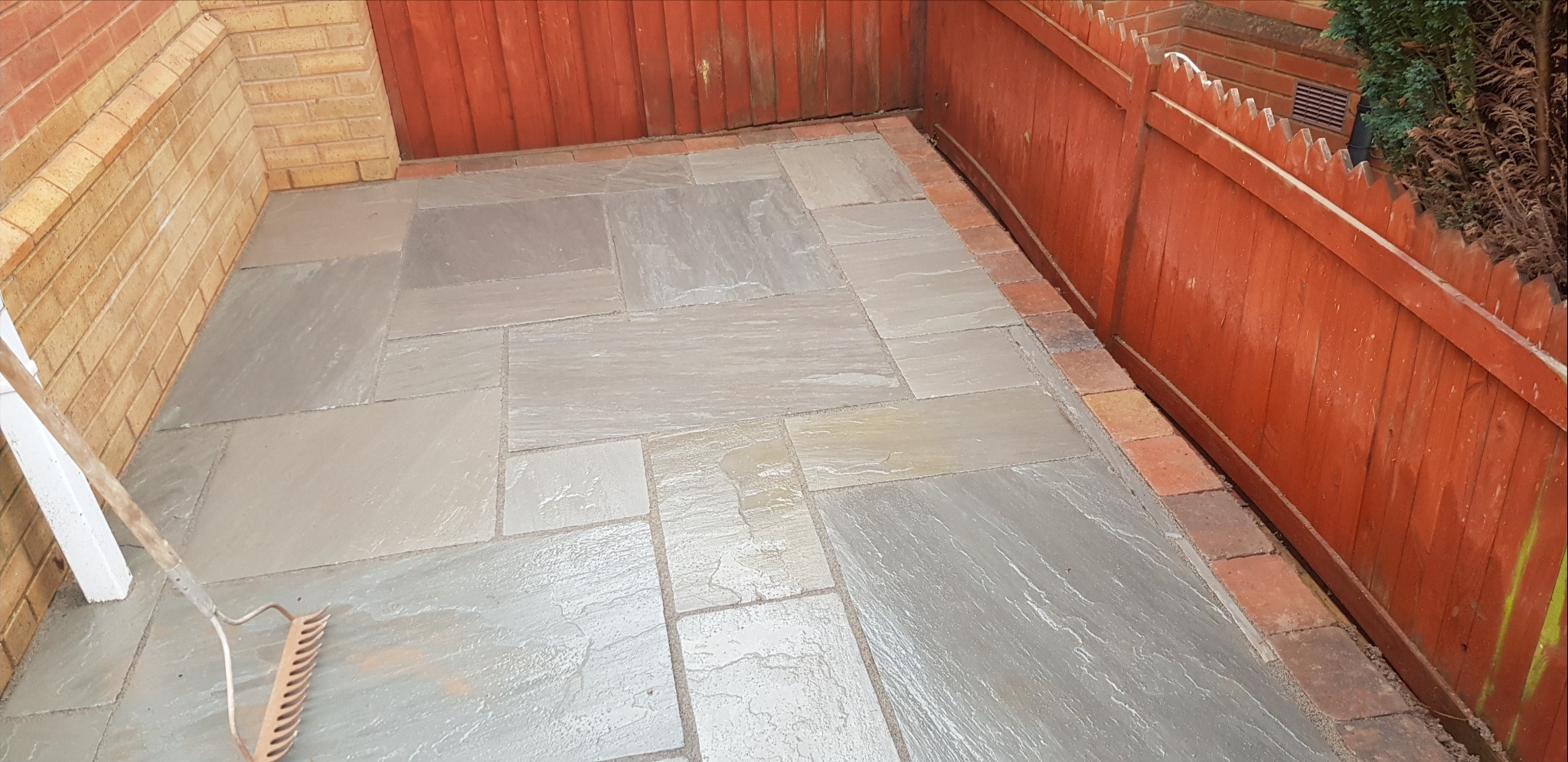 Ułożona, jeszcze mokra, szara kostka na podwórku w Olsztynie, obok drewnianego płotu pomalowanego na czerwono, z widocznymi śladami po grabiach.