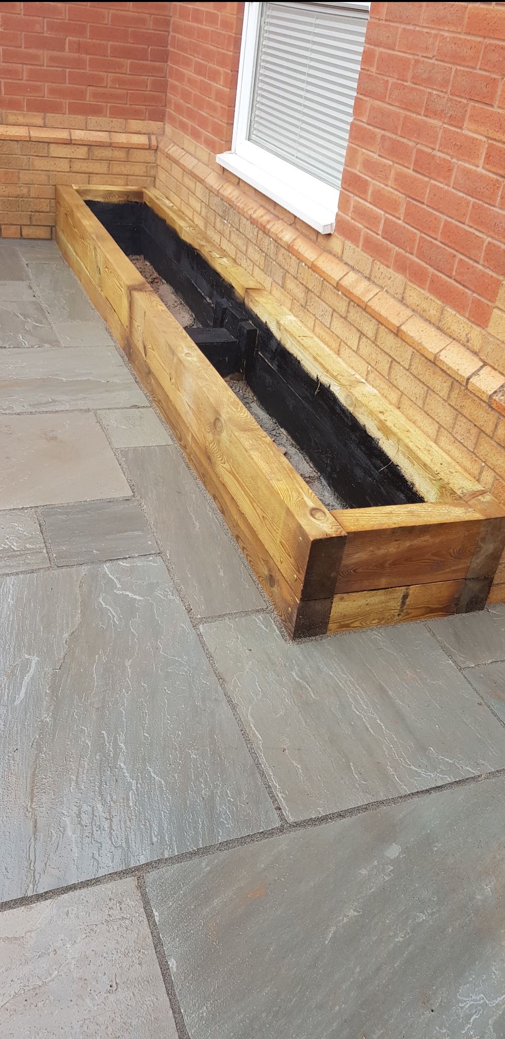 Wykonana z jasnego drewna skrzynia na rośliny, zabezpieczona czarną powłoką od wewnątrz, ustawiona na szarym kamiennym tarasie przy ceglanej ścianie budynku z oknem.