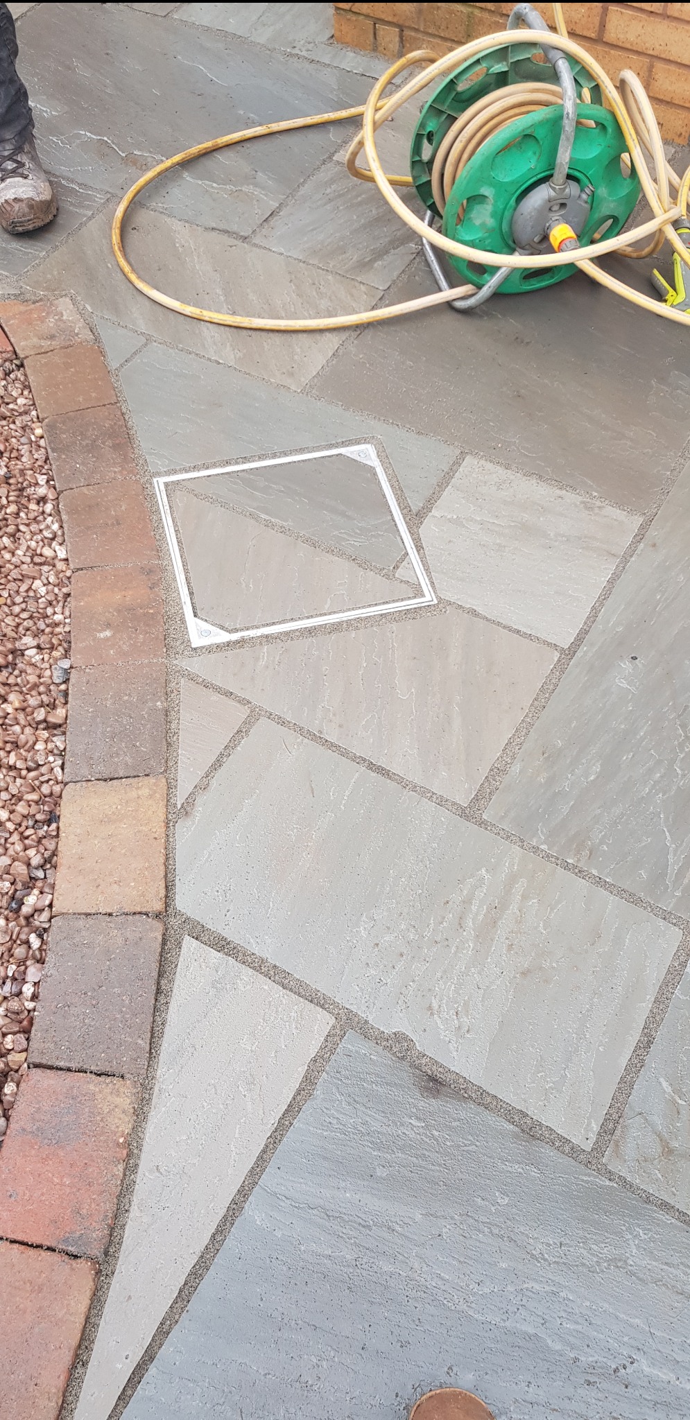 Ułożona nawierzchnia z nieregularnych płyt kamiennych w odcieniach szarości, z widoczną studzienką rewizyjną, obrzeżem z cegły i rolką węża ogrodowego na wierzchu.