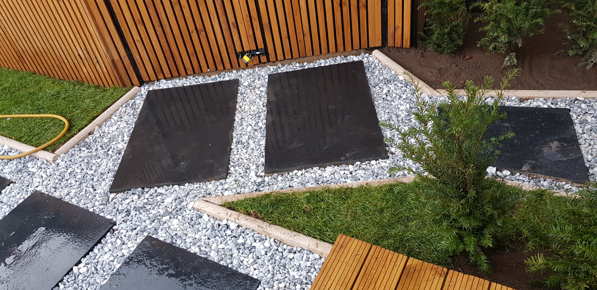 Ścieżka z ciemnych płyt ułożonych na białym żwirze, otoczona zielenią trawy i niskich krzewów, z drewnianym ogrodzeniem w tle i fragmentem drewnianego tarasu w rogu.