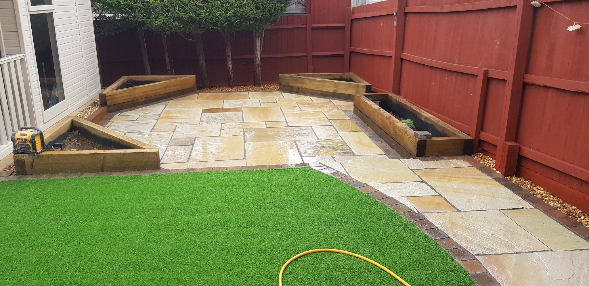 Wykończony taras z piaskowca z drewnianymi donicami w kształcie trójkątów i prostokątów, sztuczna trawa, czerwone ogrodzenie w tle.